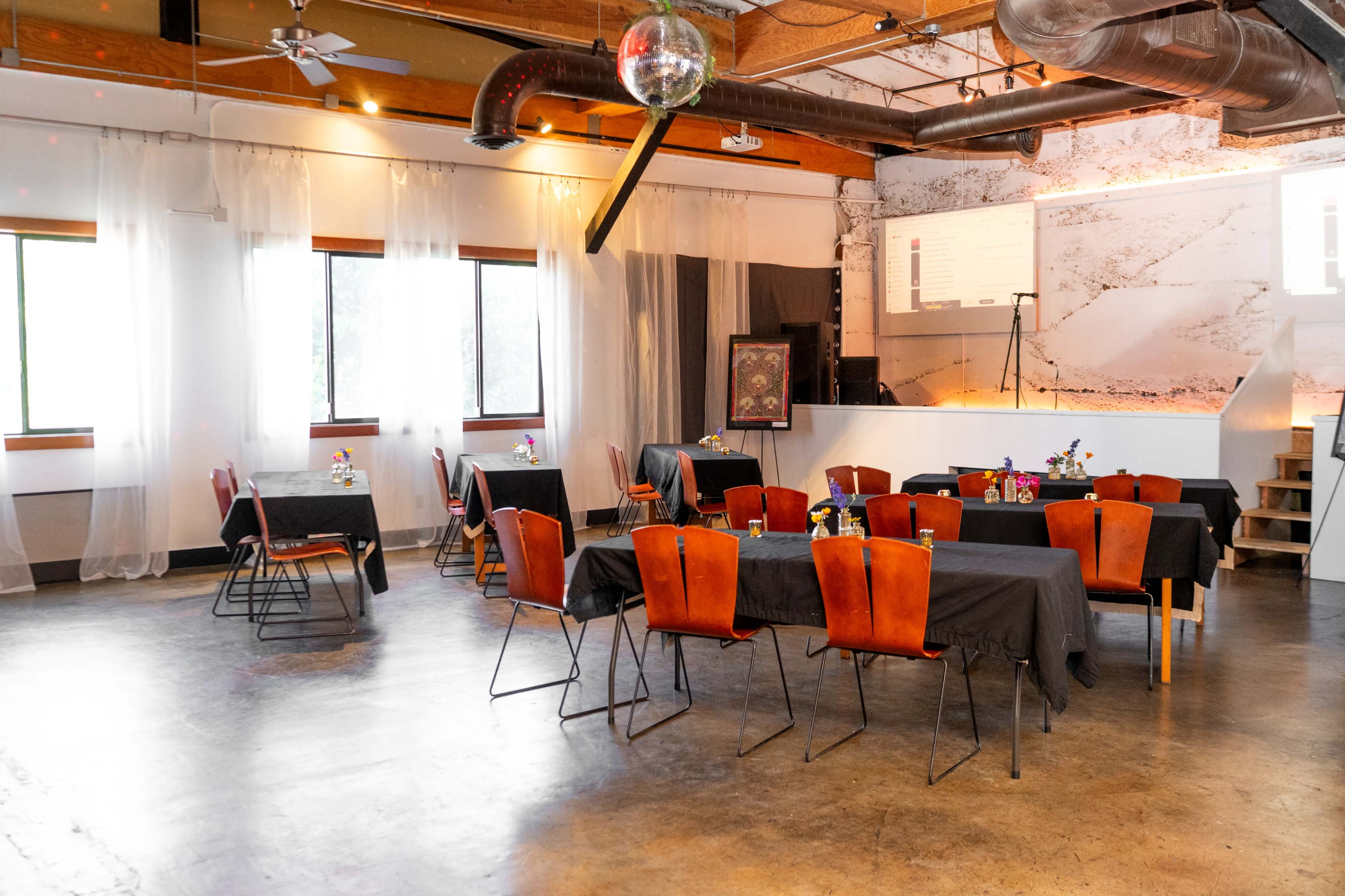 A modern event space features several tables covered with black tablecloths, arranged in a semi-circle, with decorative centerpieces and large windows allowing natural light to enter.