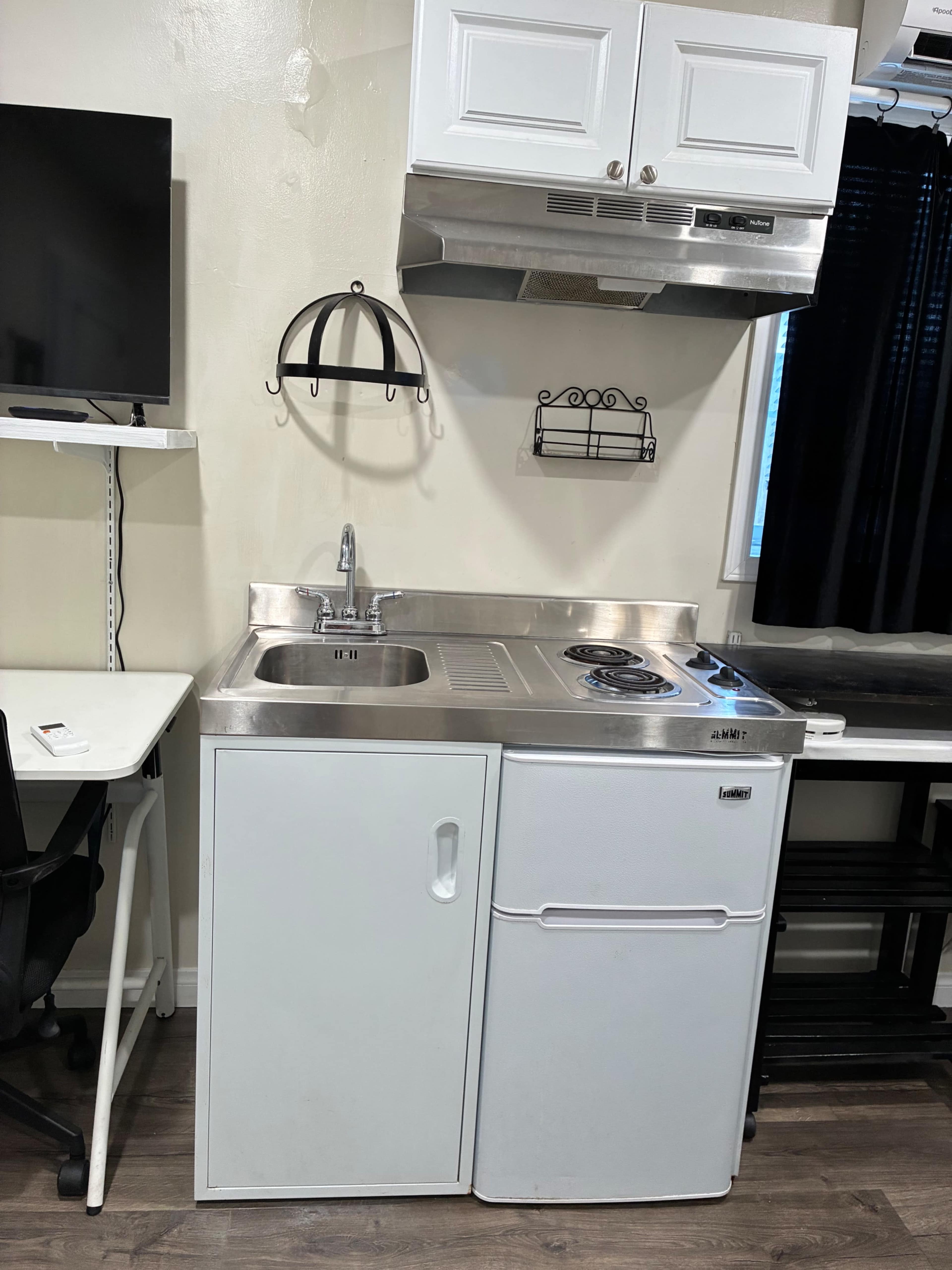 The image shows a small kitchen area featuring a stainless steel sink, a two-burner electric stove, a white refrigerator, and a wall-mounted shelf, all within a compact space.