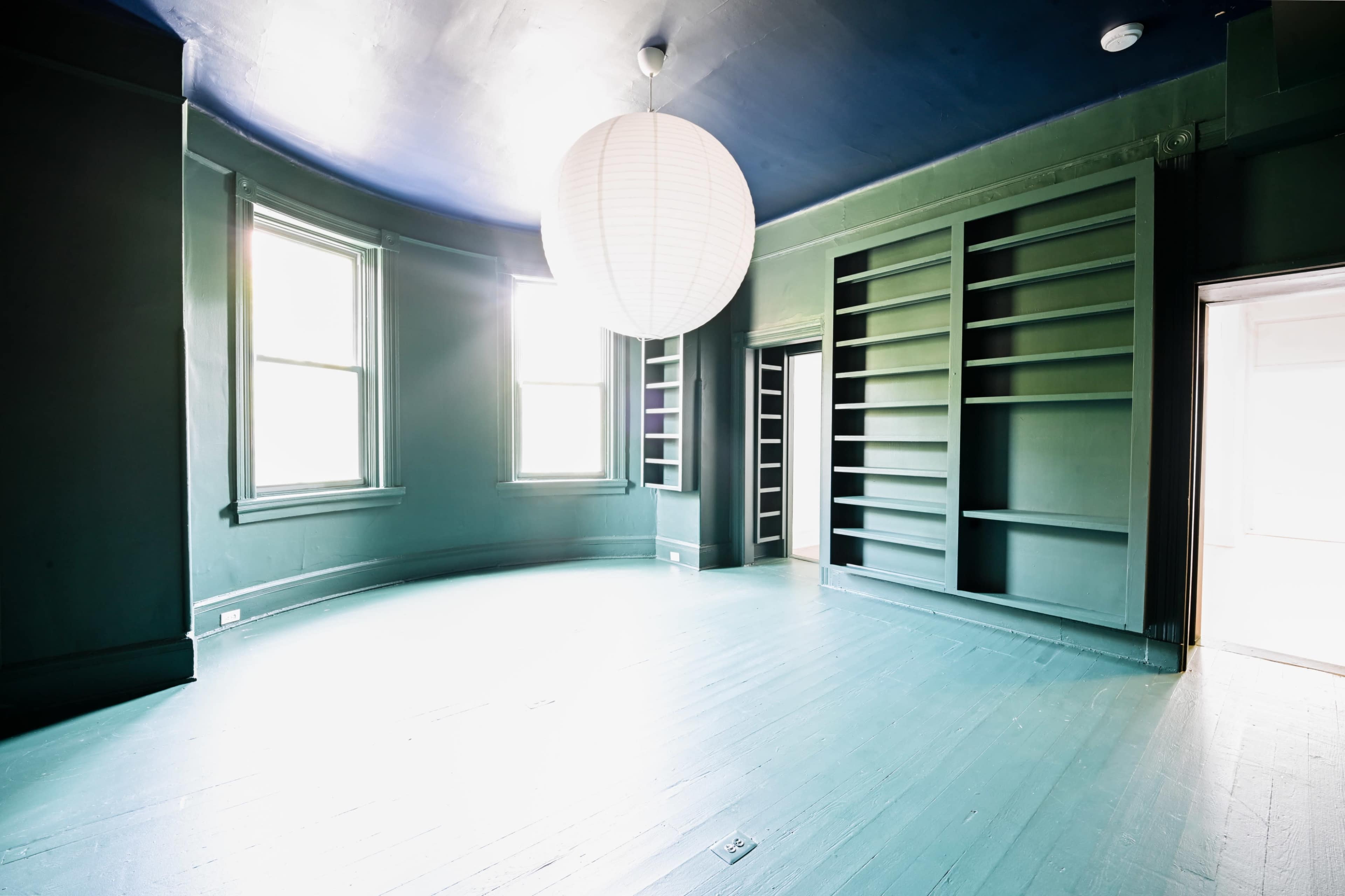 The image shows a spacious room with green walls, large windows, and a minimalist design featuring a round pendant light and built-in shelves.