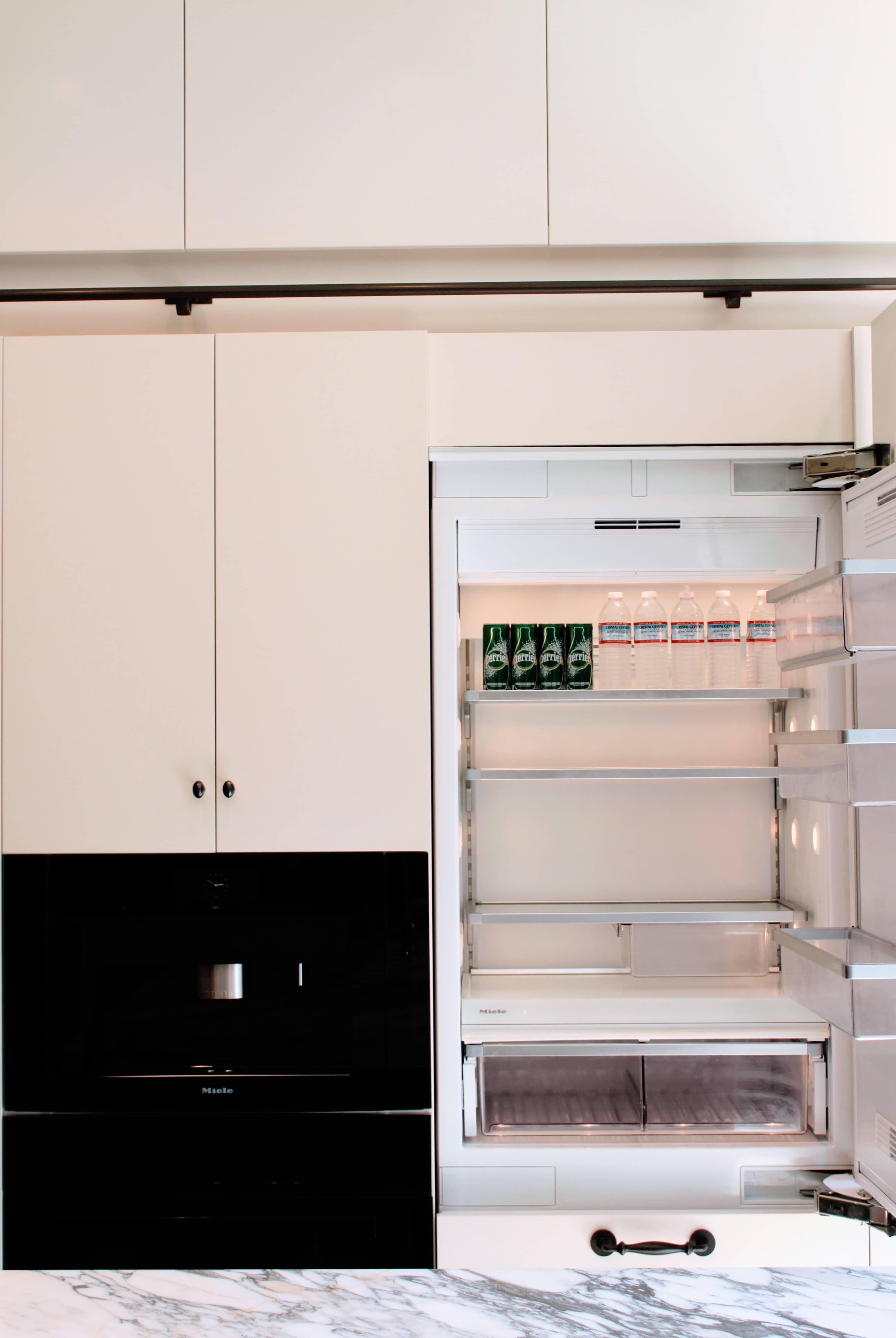 A modern kitchen featuring an open refrigerator with glass shelves and several bottles of water inside, alongside sleek cabinetry and a built-in microwave.