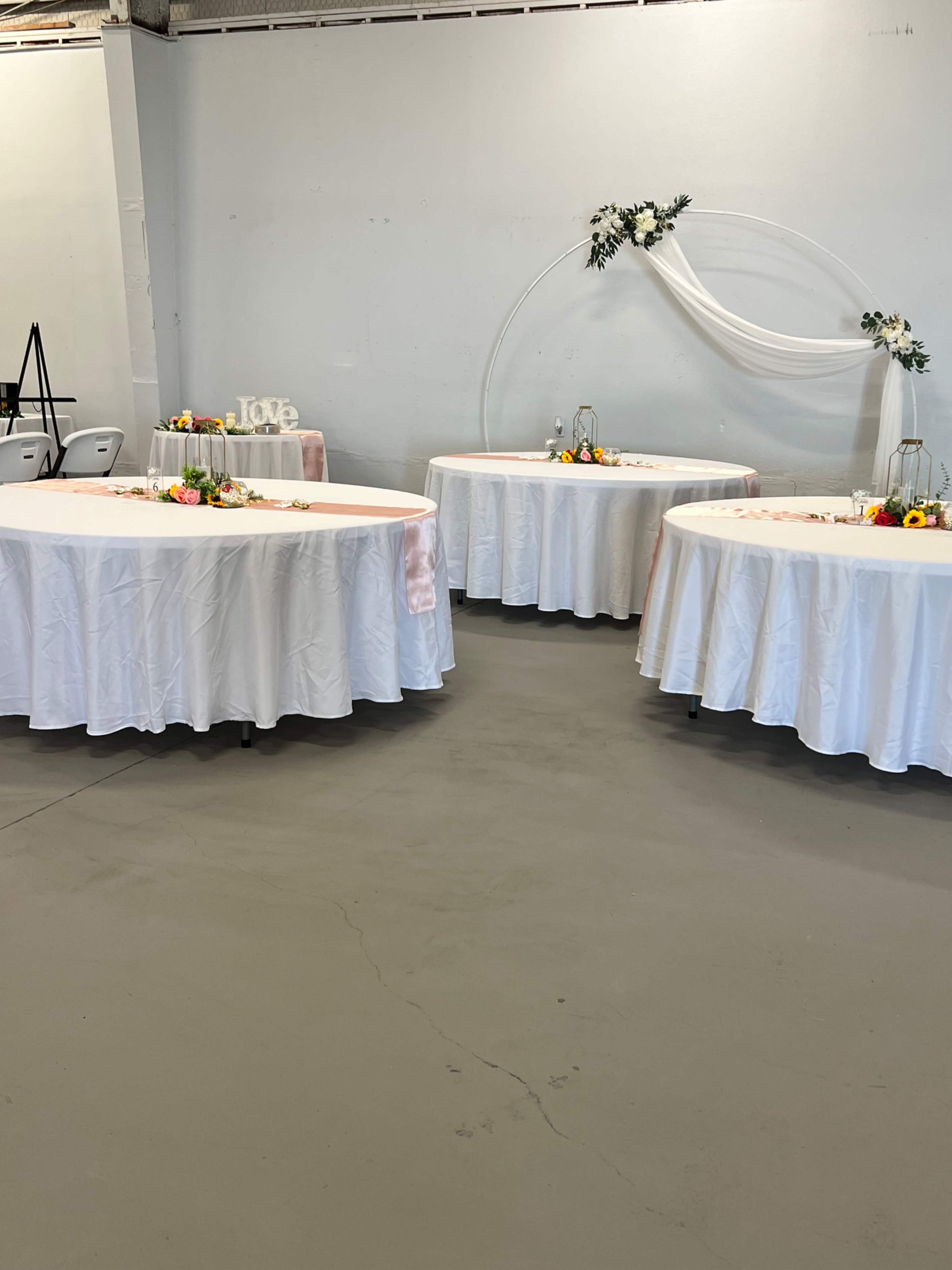The image shows a venue with three round tables covered in white tablecloths, adorned with floral centerpieces and a decorative backdrop featuring drapery and a circular arrangement.