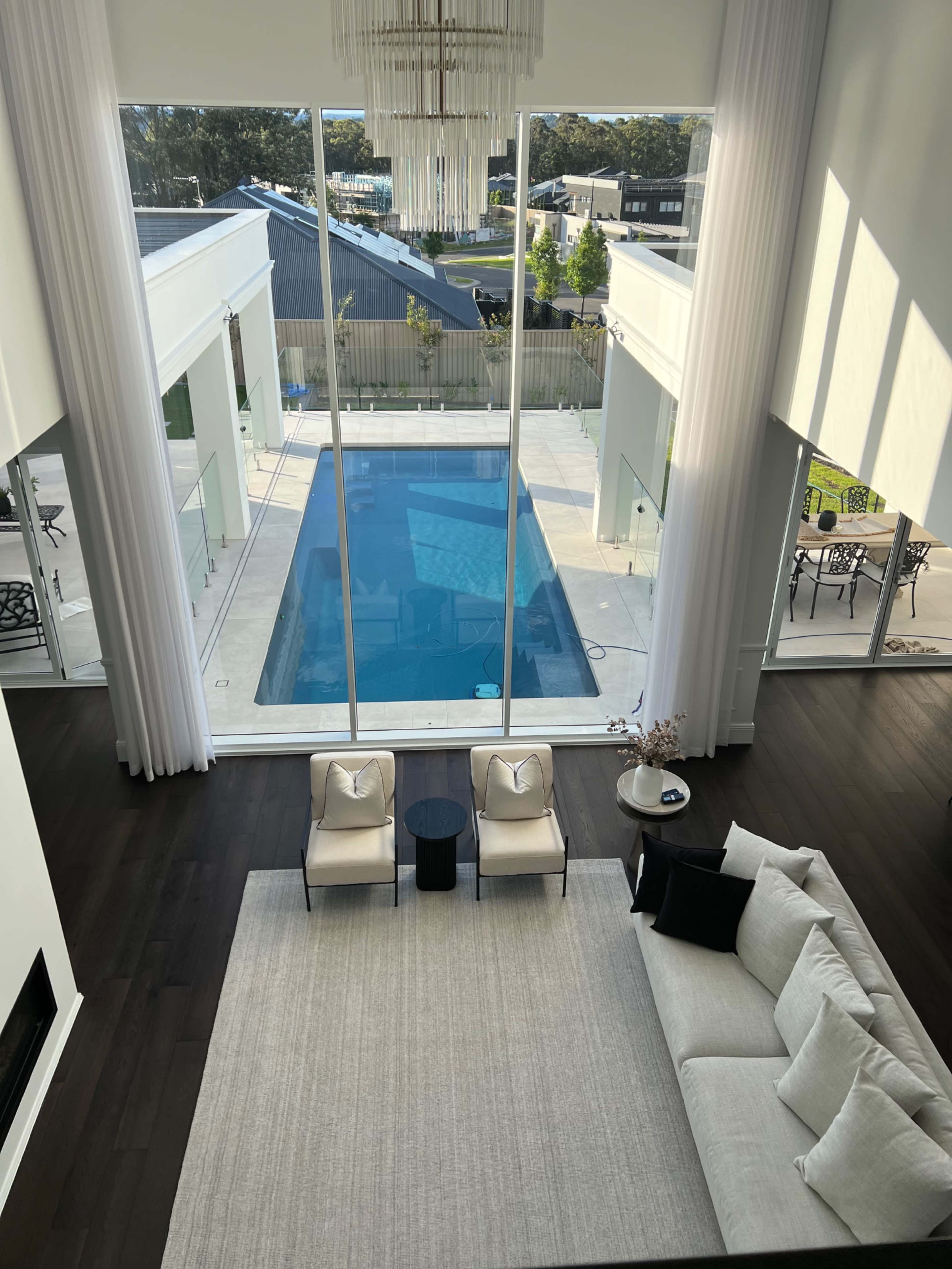 The image shows a modern living room overlooking a swimming pool through large glass windows, with minimalist furniture and a neutral color palette.