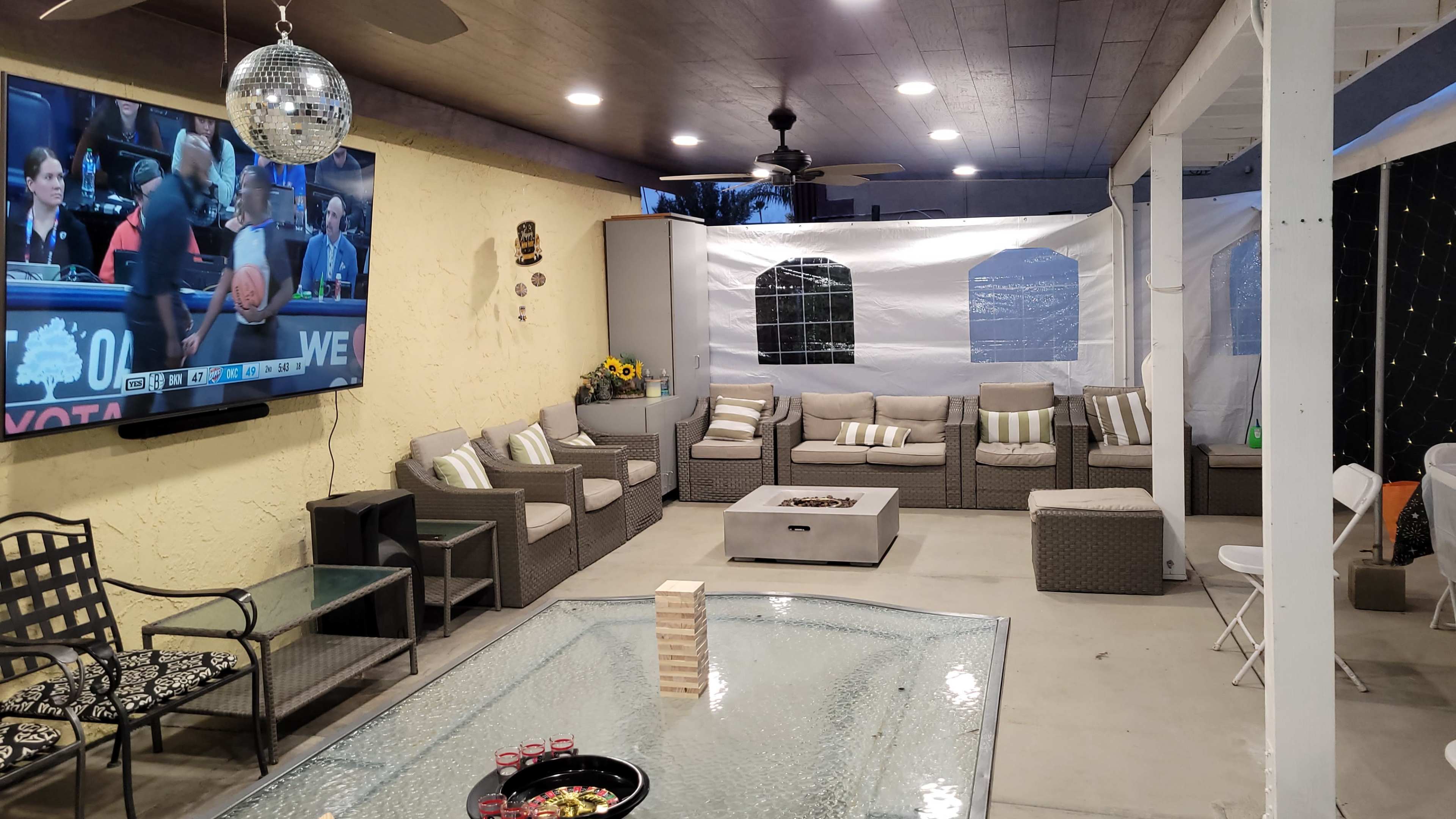 A covered outdoor living space with a TV, seating area, and a glass-topped table.