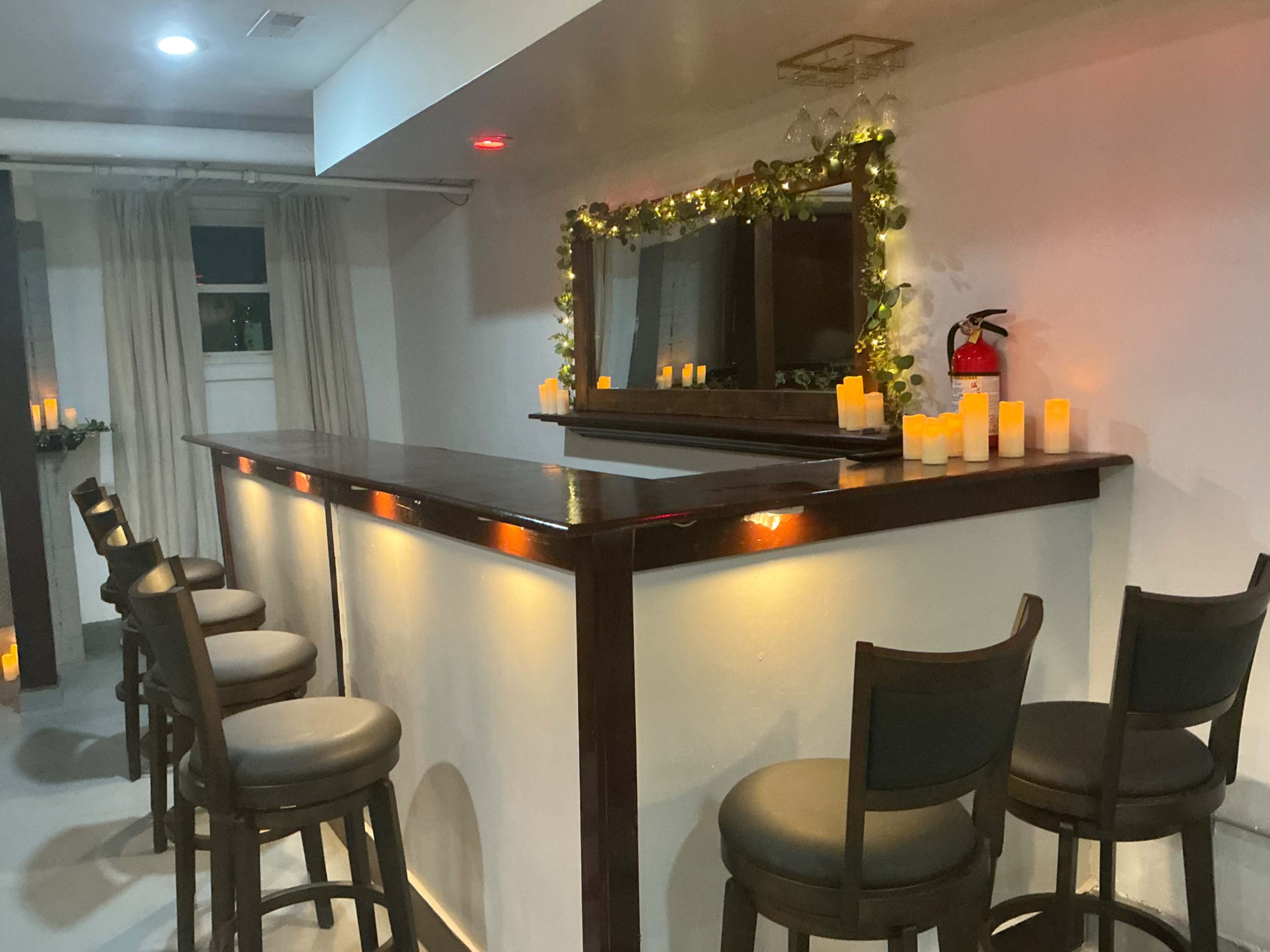 The image shows a dimly lit bar area with a long wooden counter, high stools, and flickering candles arranged on the bar and around the space.