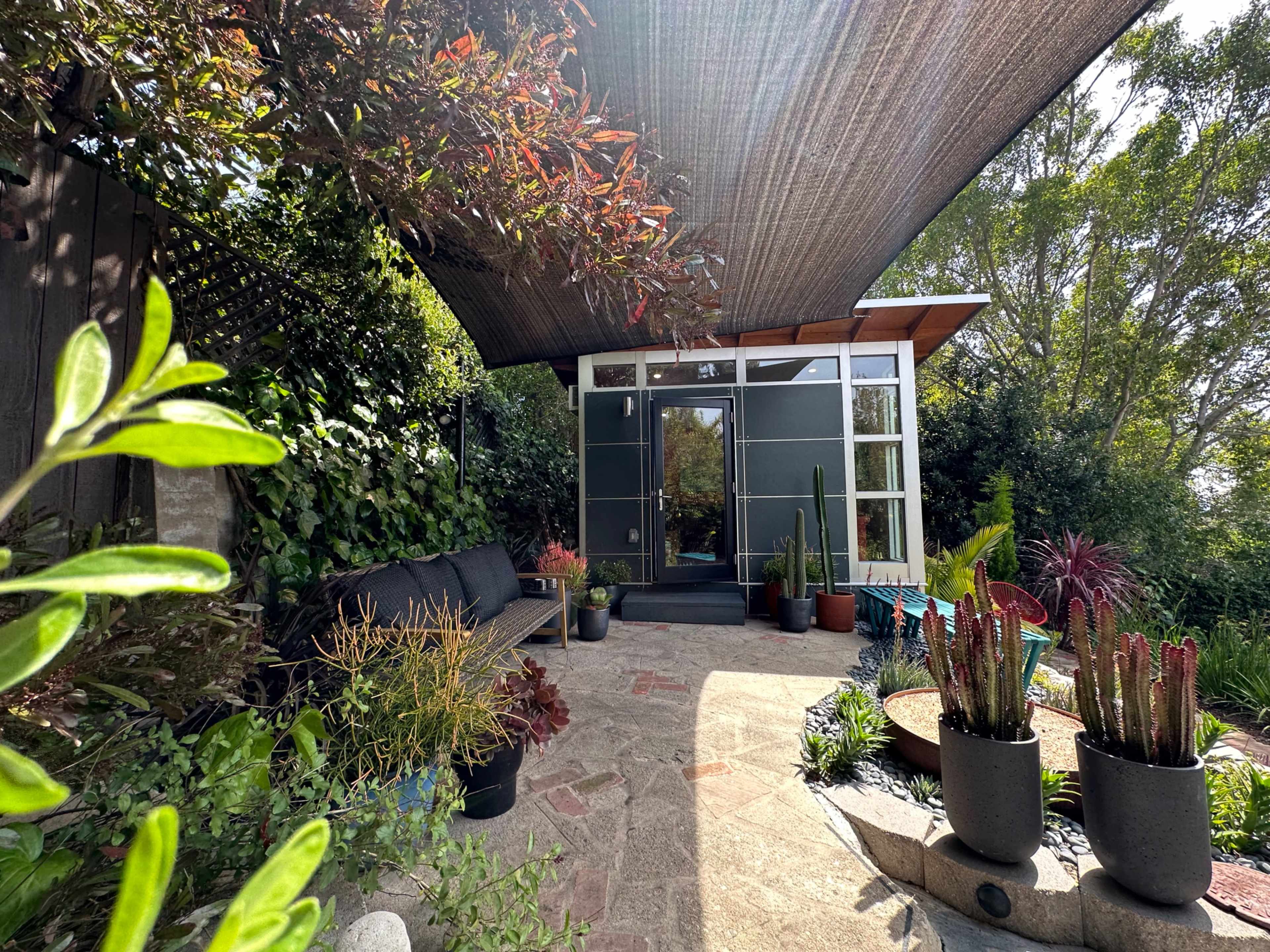 A modern garden area features a seating arrangement surrounded by various potted plants and a shaded entrance.