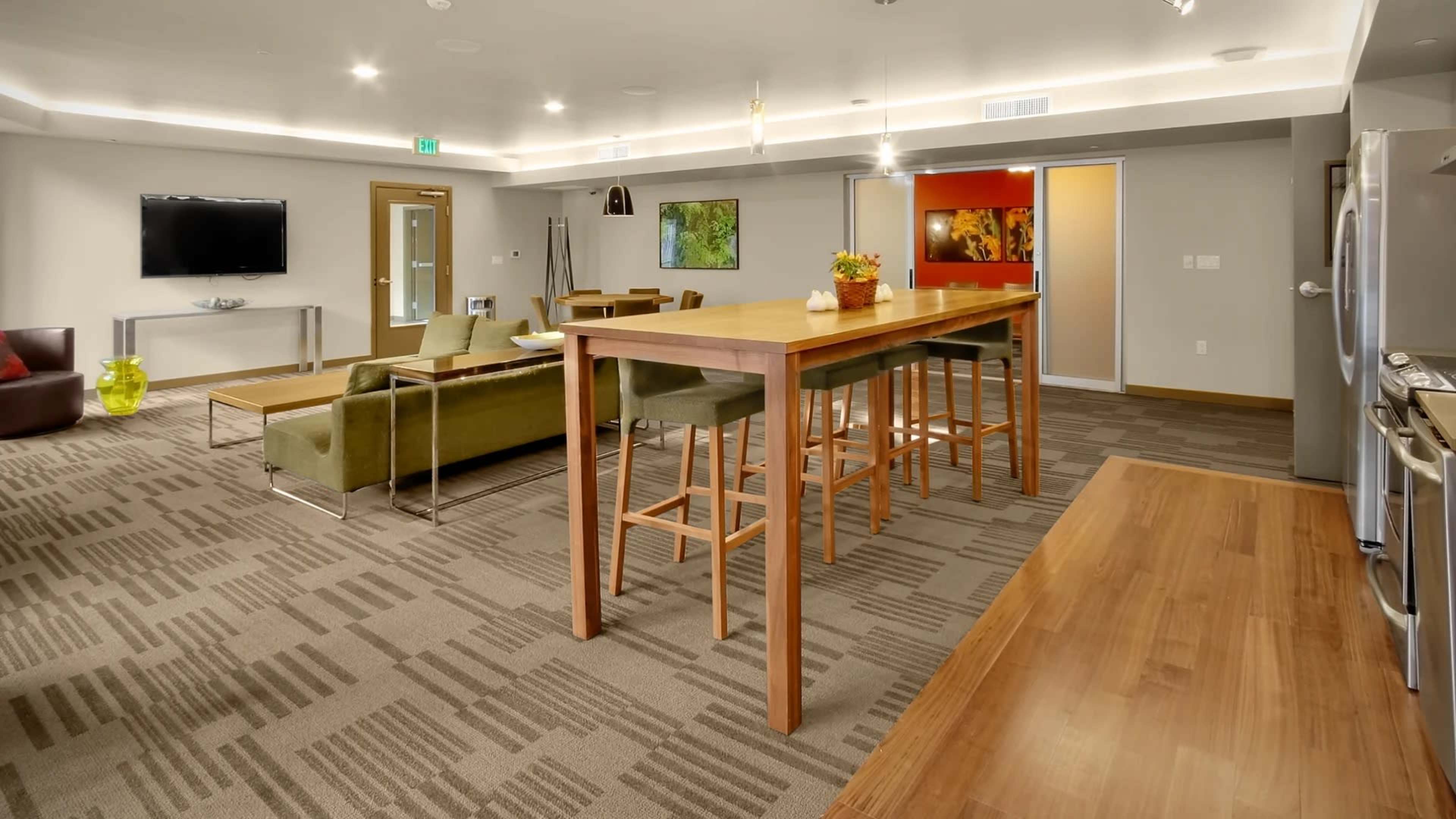A modern common area featuring a large wooden table surrounded by high stools, a green couch, and a television on the wall.