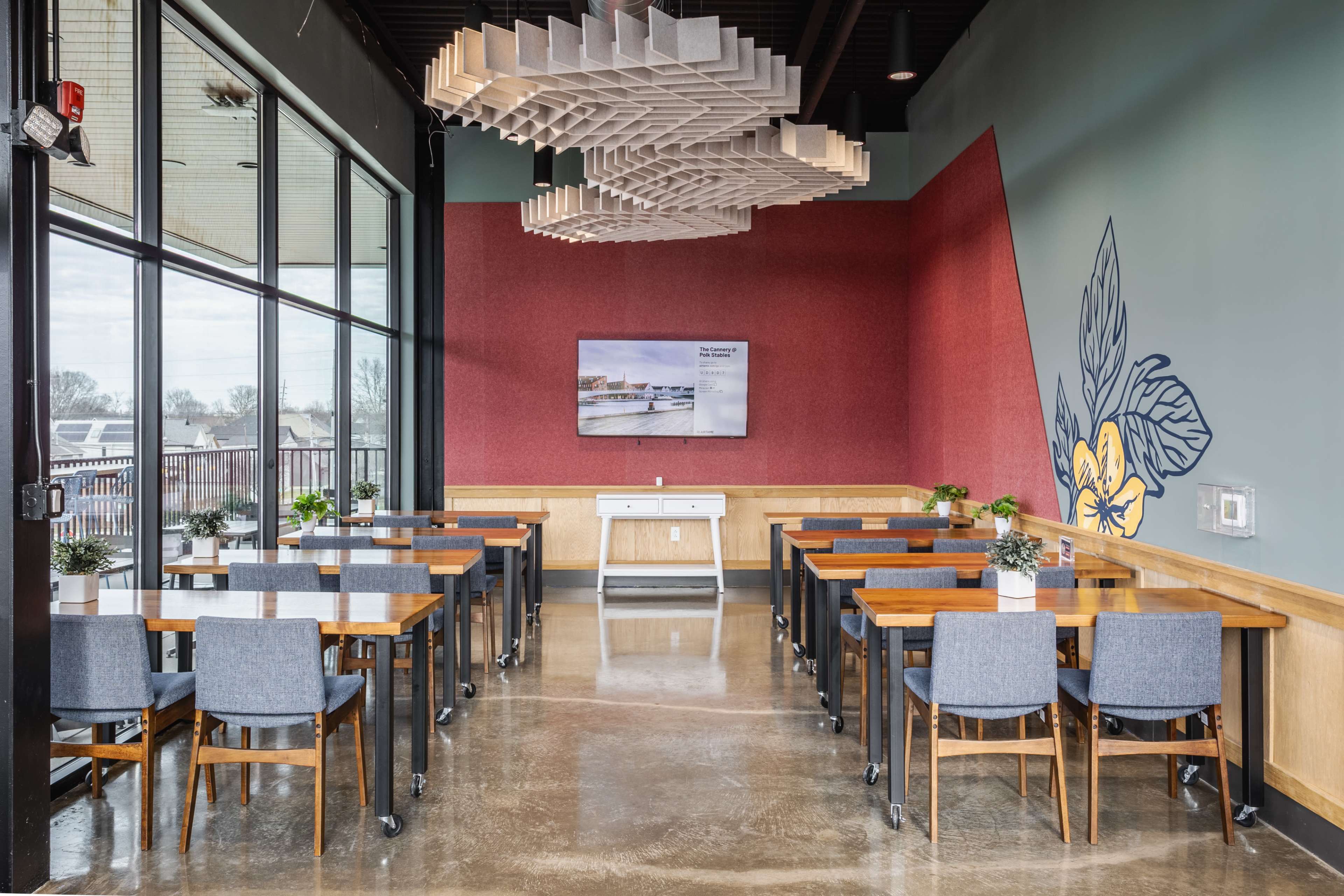 The image shows a modern dining area with multiple tables and chairs, large windows allowing natural light, and decorative acoustic panels on the ceiling.