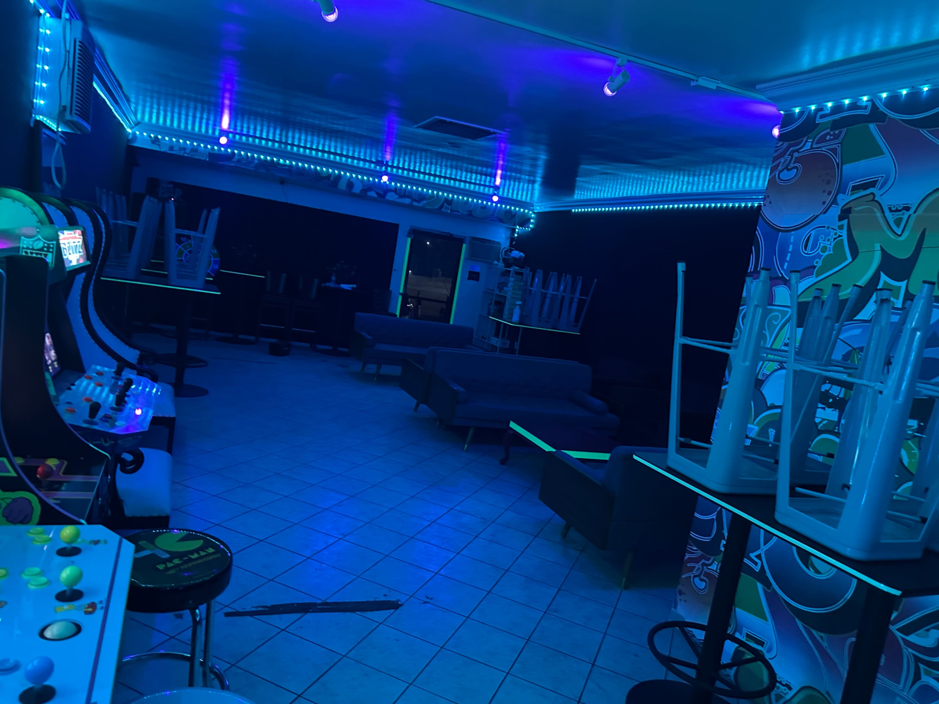 The image shows a vacant arcade room with neon blue lighting, featuring empty game machines and tables stacked upside down.