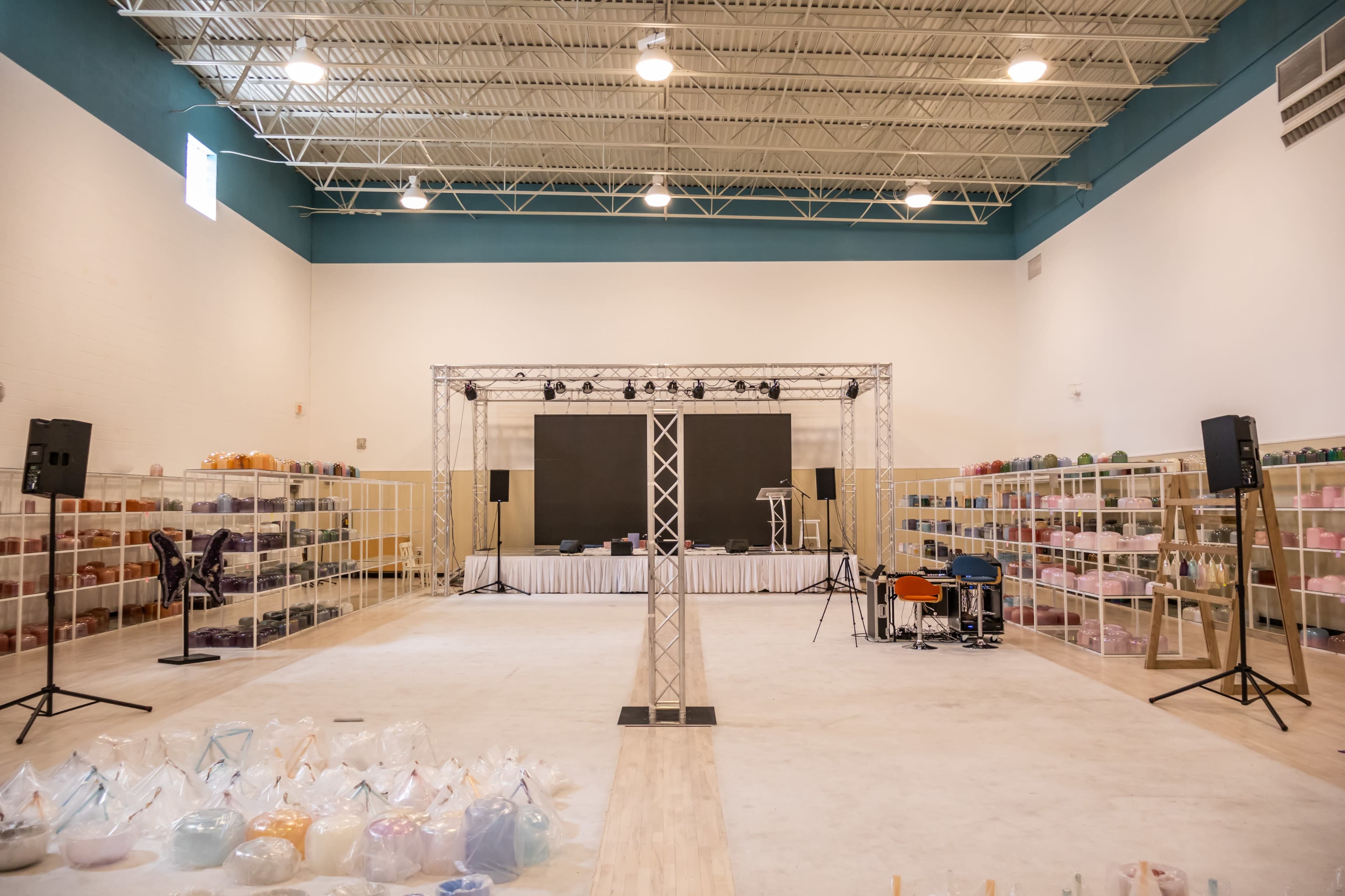 The image shows a spacious interior of a room set up for an event, featuring a stage at one end and shelves filled with colorful containers along the walls.
