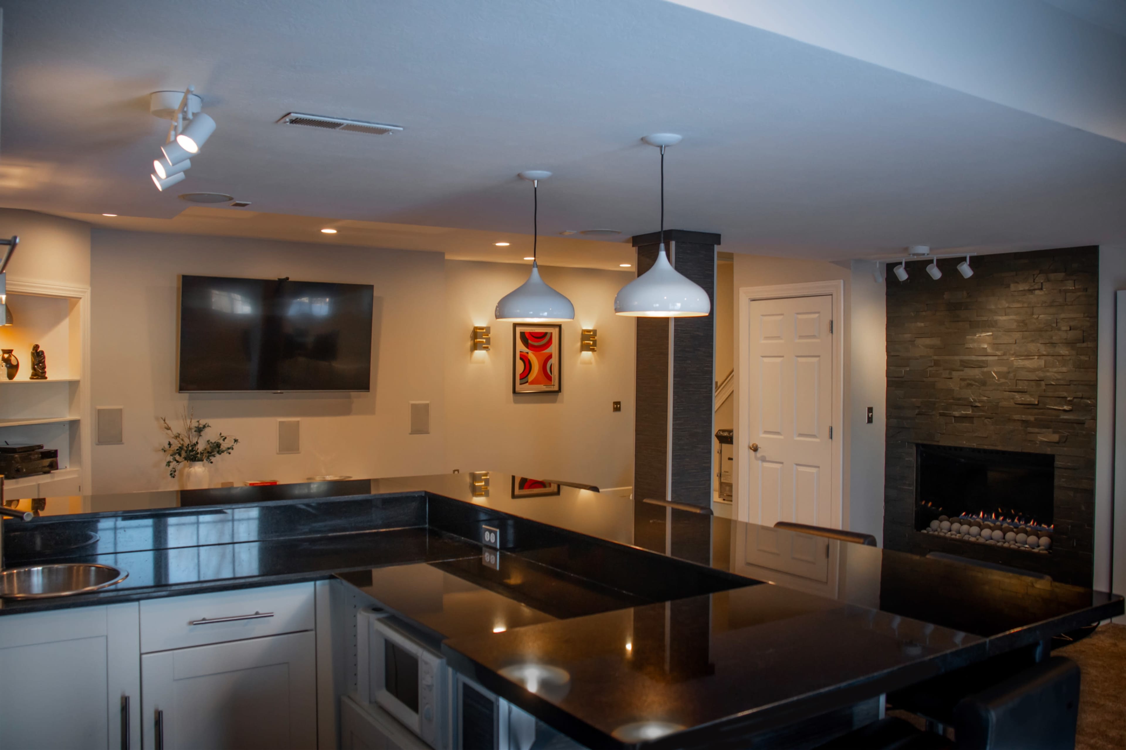 The image shows a modern kitchen with a large island, pendant lighting, a wall-mounted television, and a fireplace in an adjacent area.