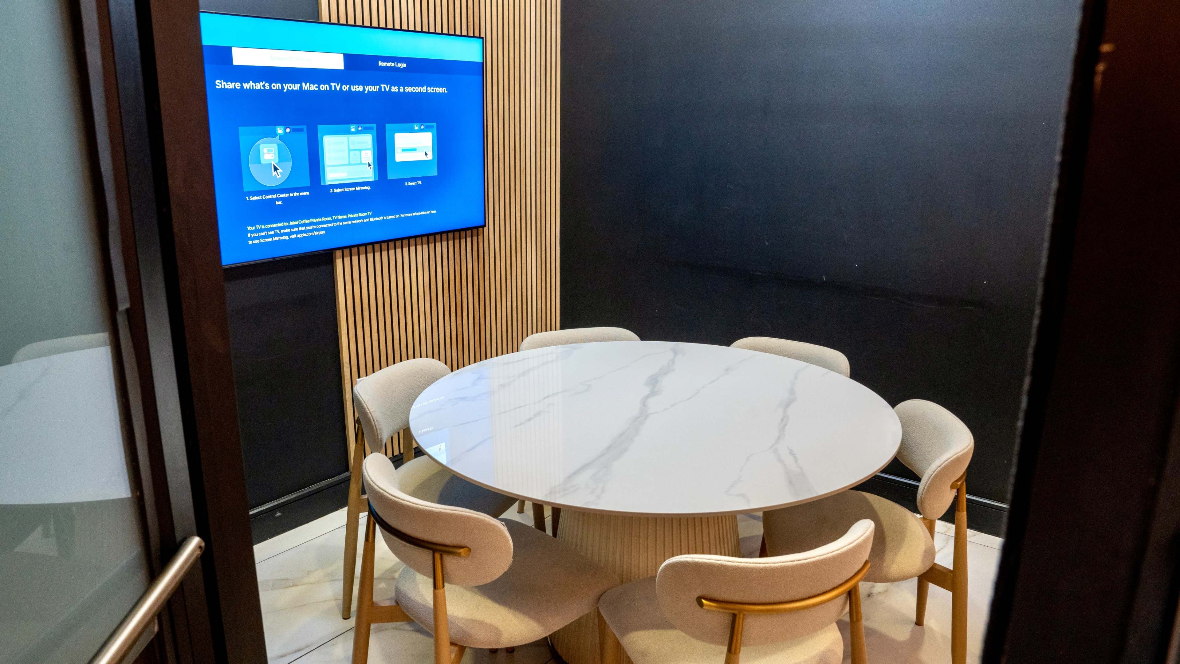 A small conference room features a round marble table surrounded by six beige chairs, with a screen displaying content on a dark wall.