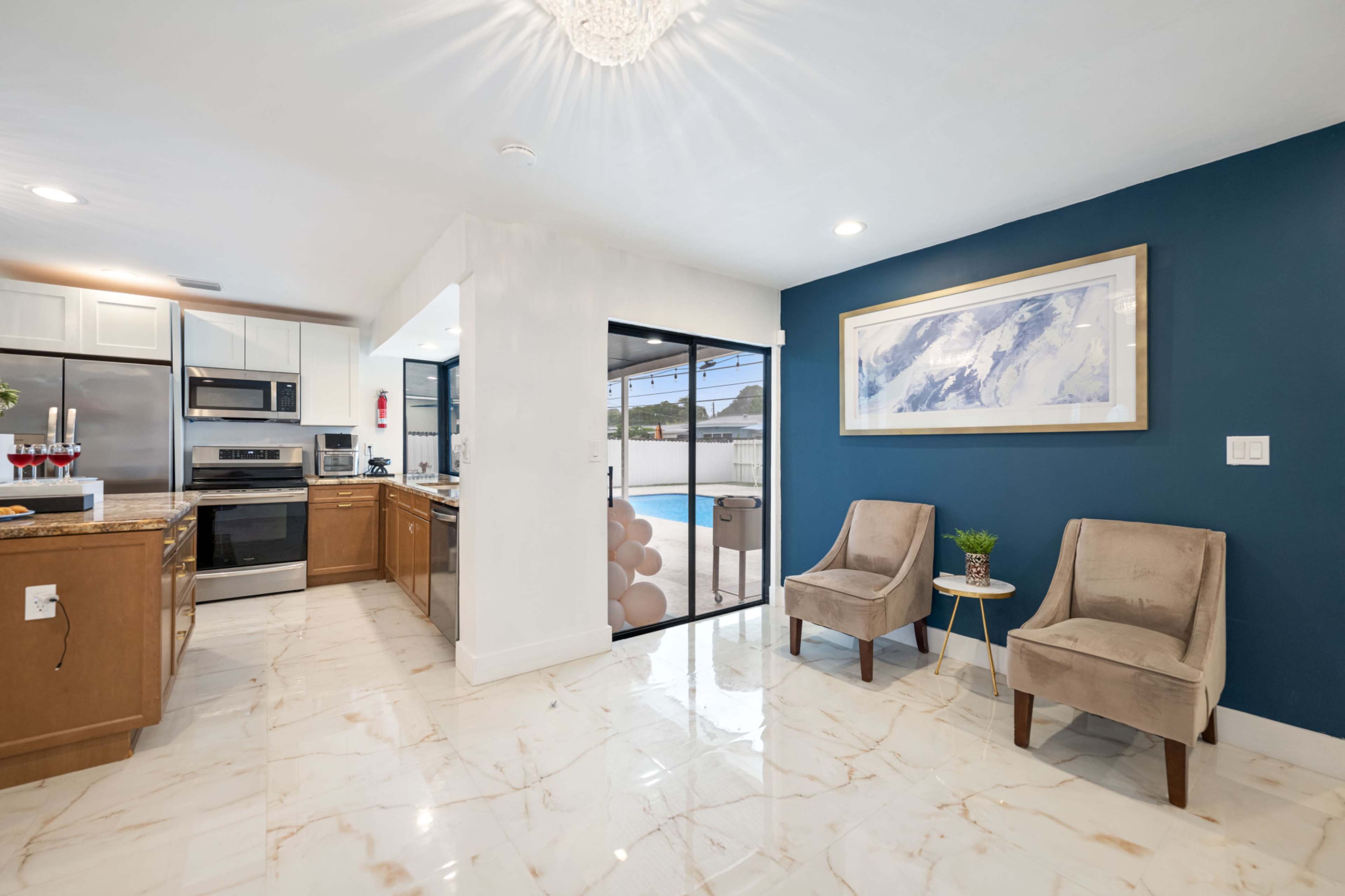 The image shows a modern kitchen with stainless steel appliances, a granite countertop, and two beige armchairs in front of a glass door leading to an outdoor space.