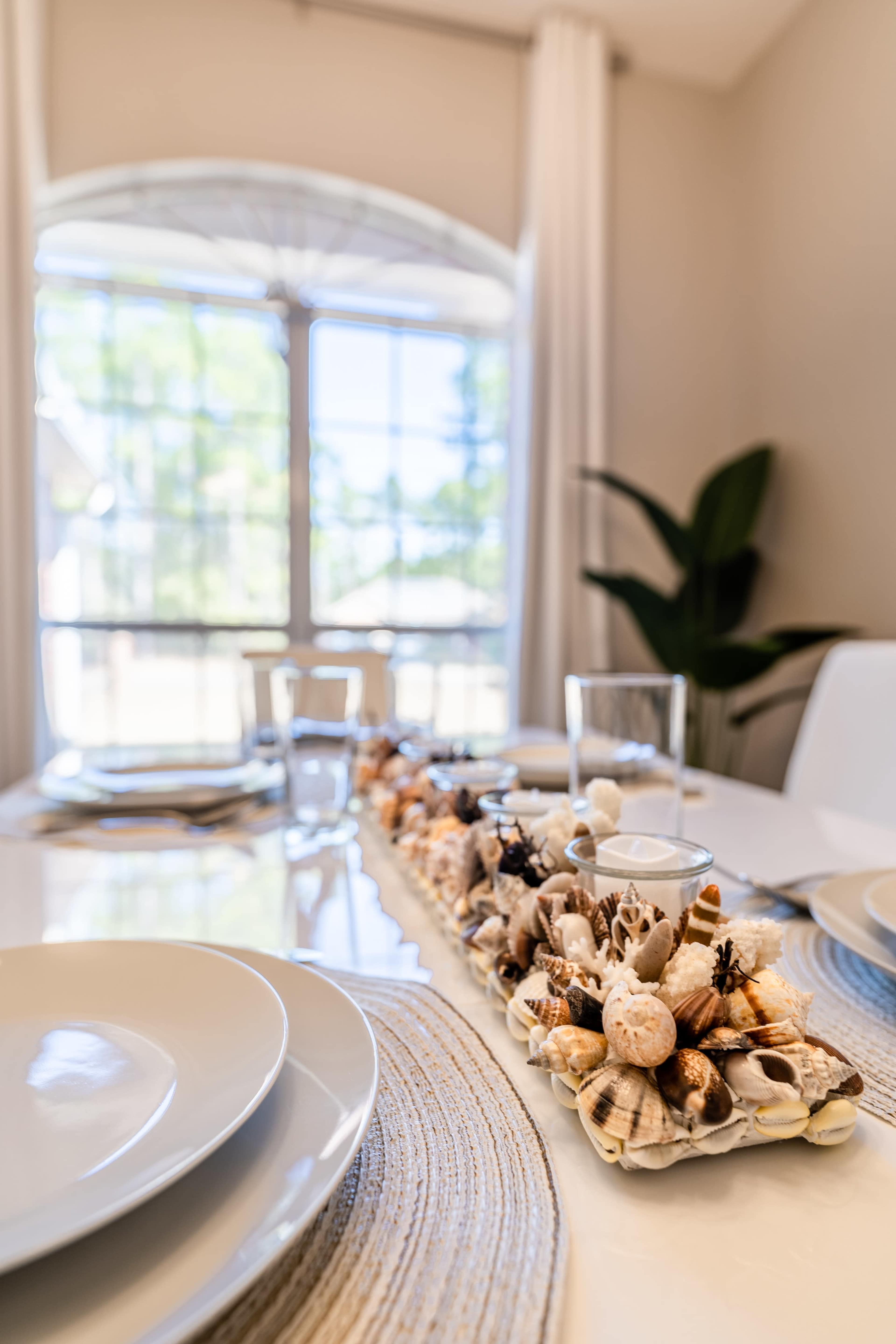 A dining table set with plates and a decorative centerpiece made of seashells, with large windows allowing natural light to enter.