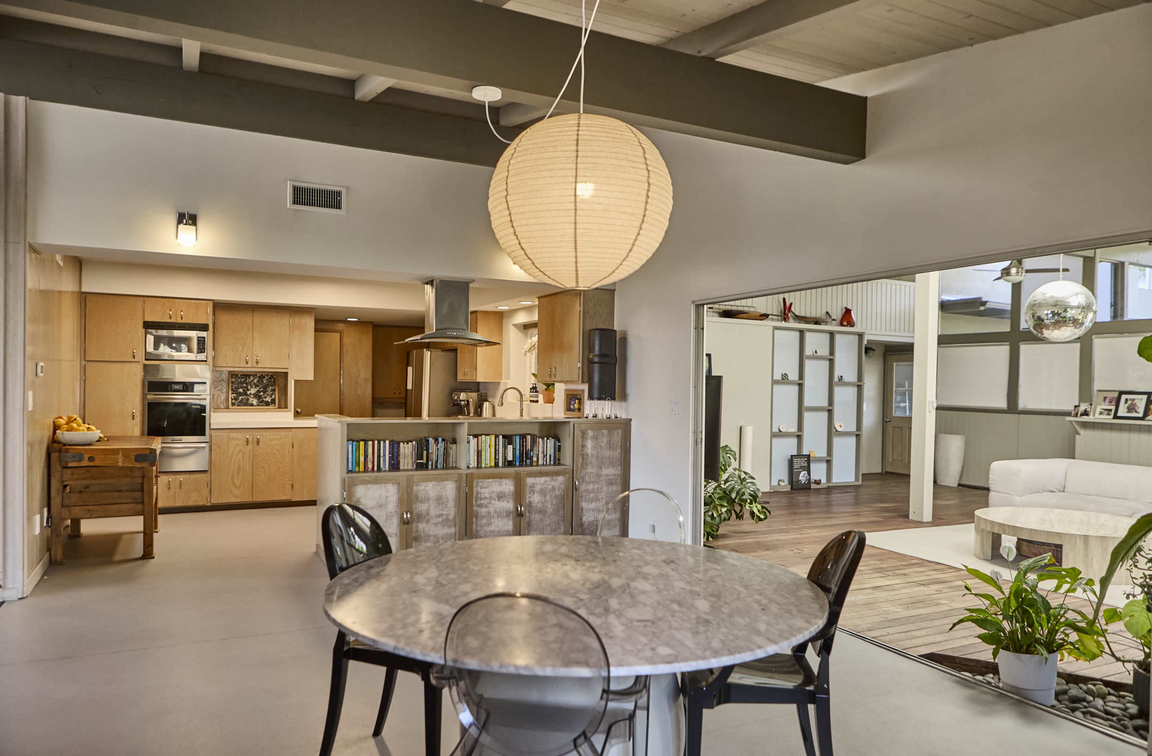 The image shows a modern, open-concept interior featuring a dining area with a round table, a kitchen in the background, and a living space with a large window overlooking a patio.