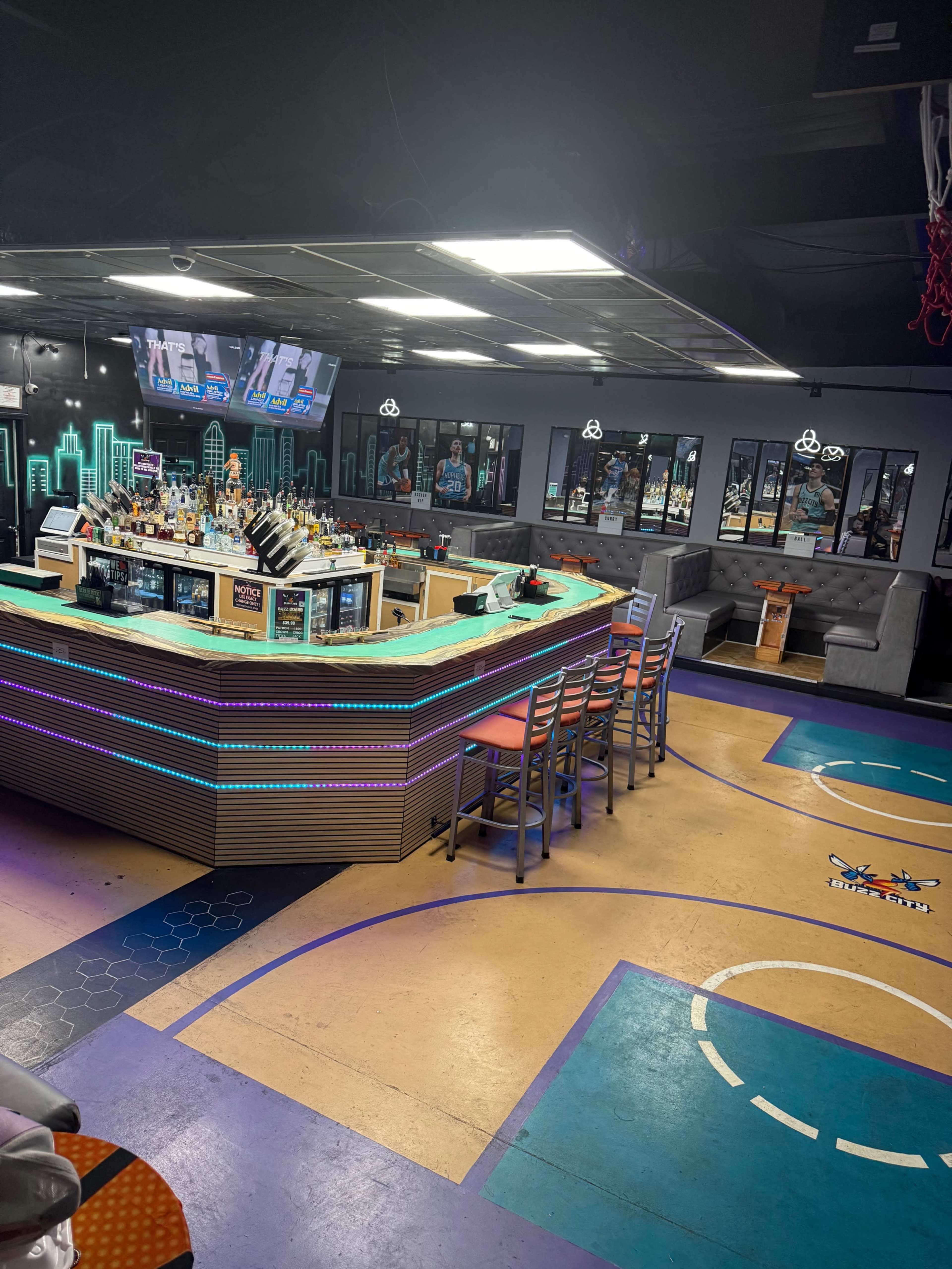 The image shows a modern bar area featuring a curved counter, colorful lighting, and sports-themed decor, with seating and monitors displaying sports content in the background.