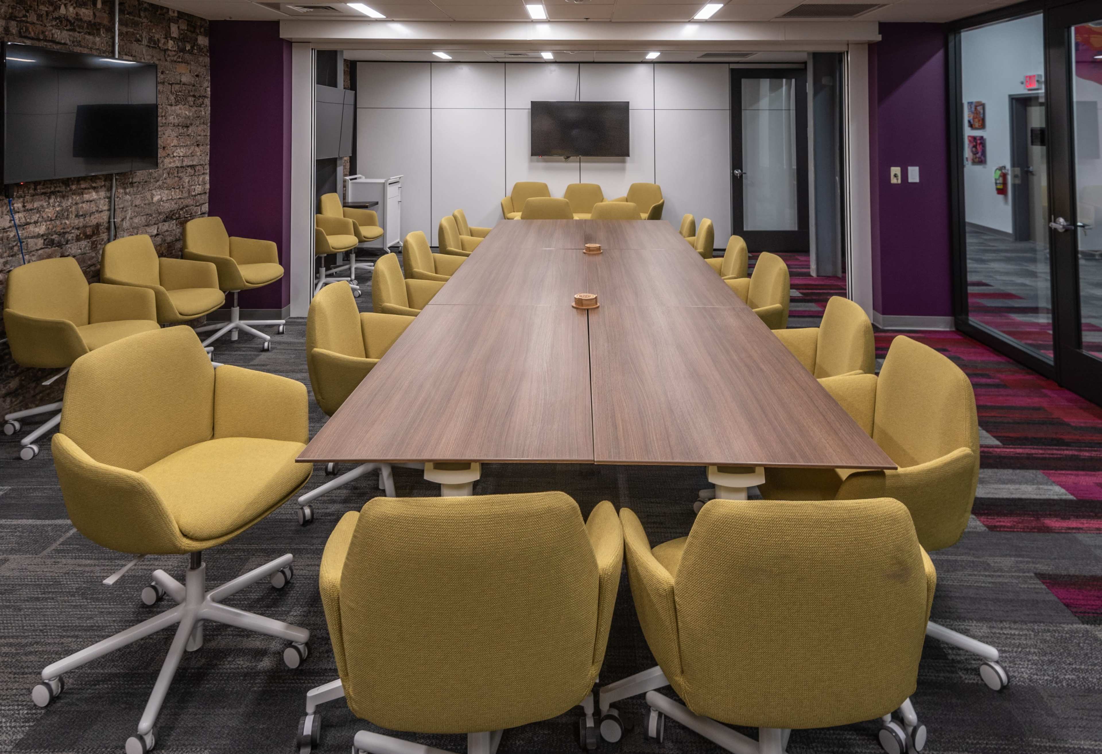 The image shows a conference room with a long wooden table surrounded by yellow swivel chairs.