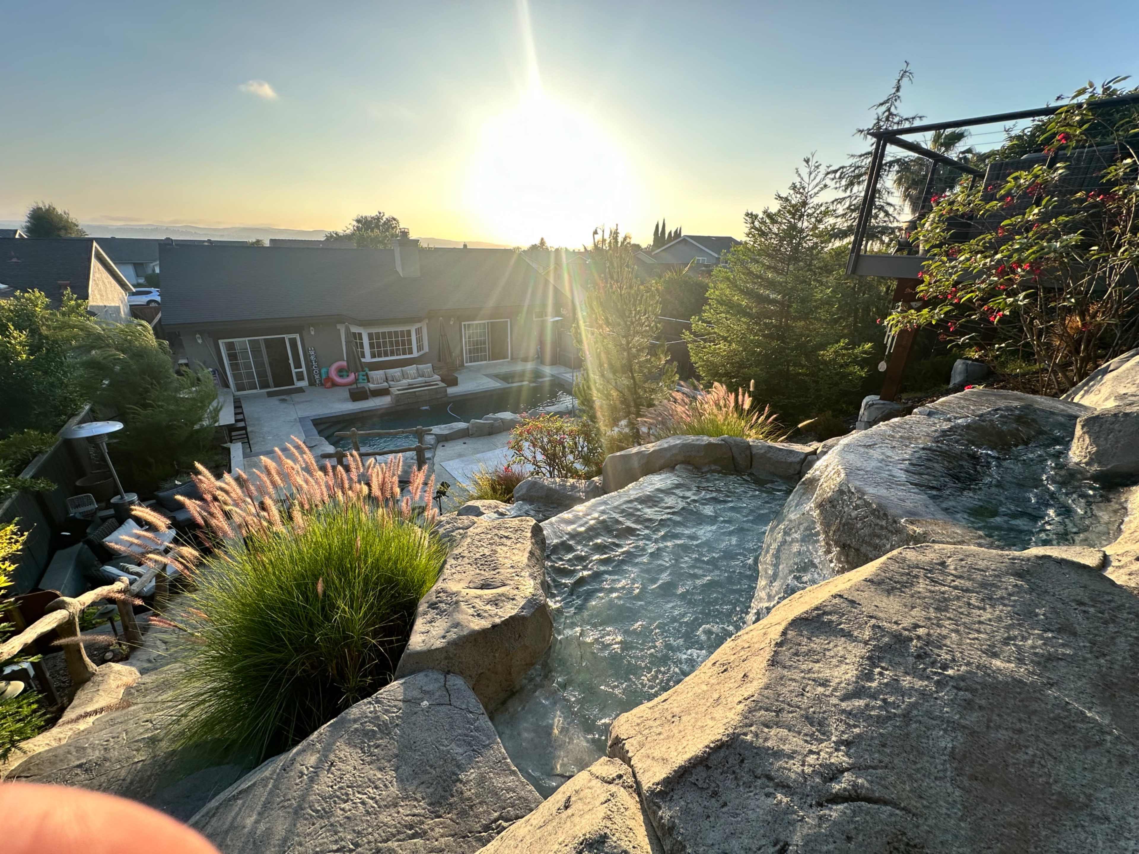 A stone hot tub and landscaping with a view of a house and a swimming pool illuminated by the sunset.