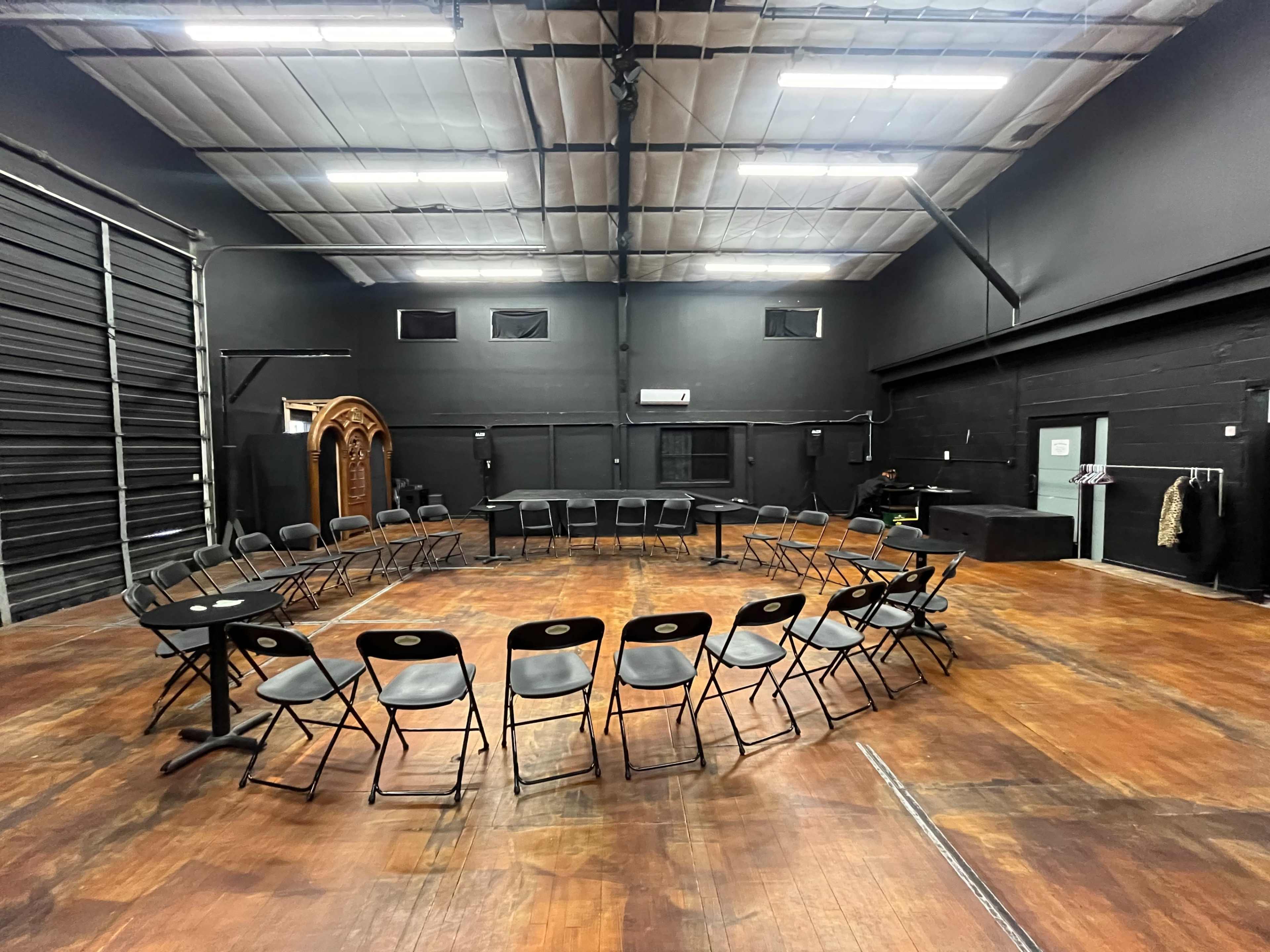 A circular arrangement of black folding chairs is set up in a dimly lit rehearsal space with black walls and wooden flooring.