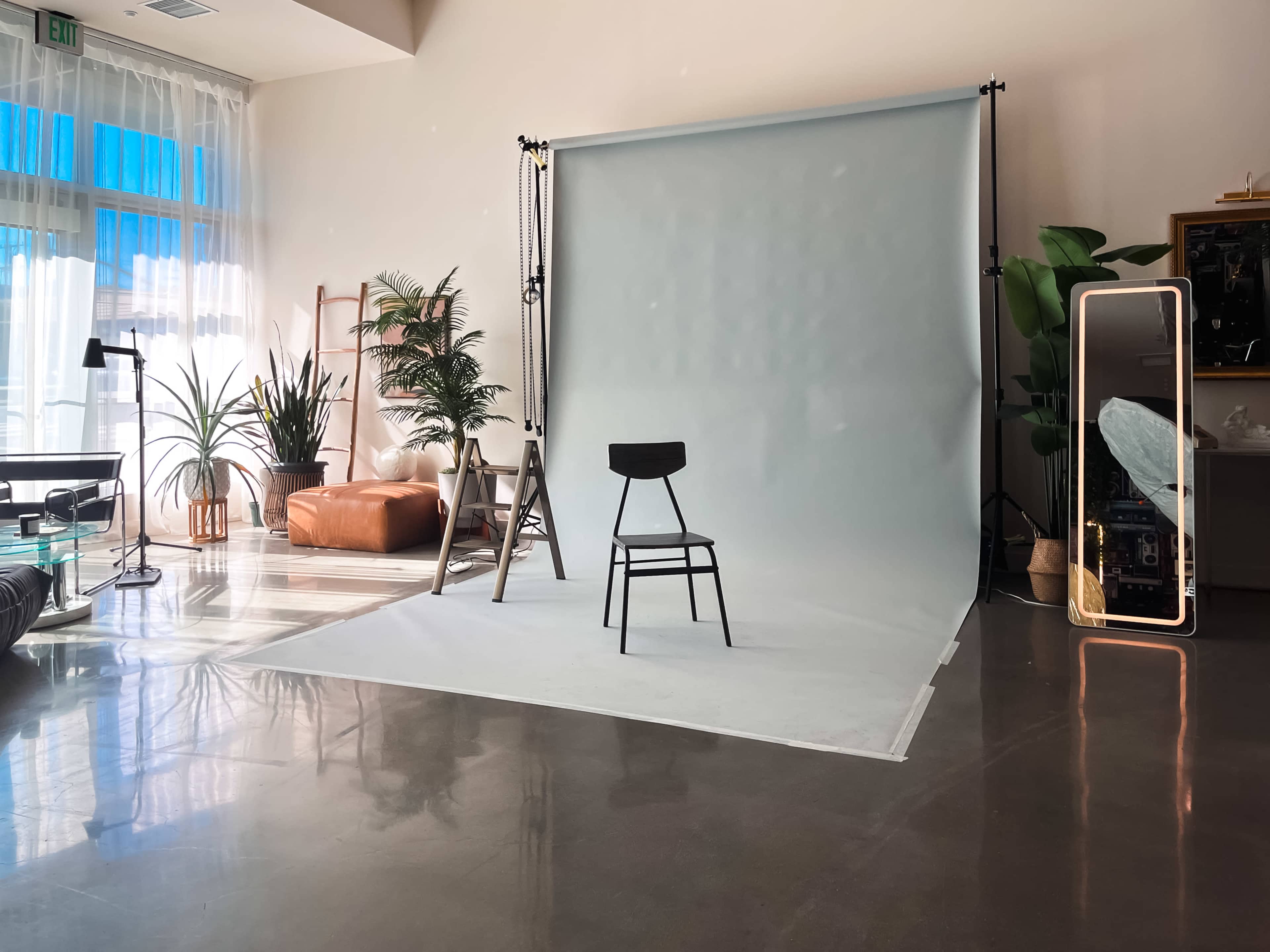 The image shows a well-lit photo studio featuring a black stool in front of a light blue backdrop, surrounded by plants and a full-length mirror.