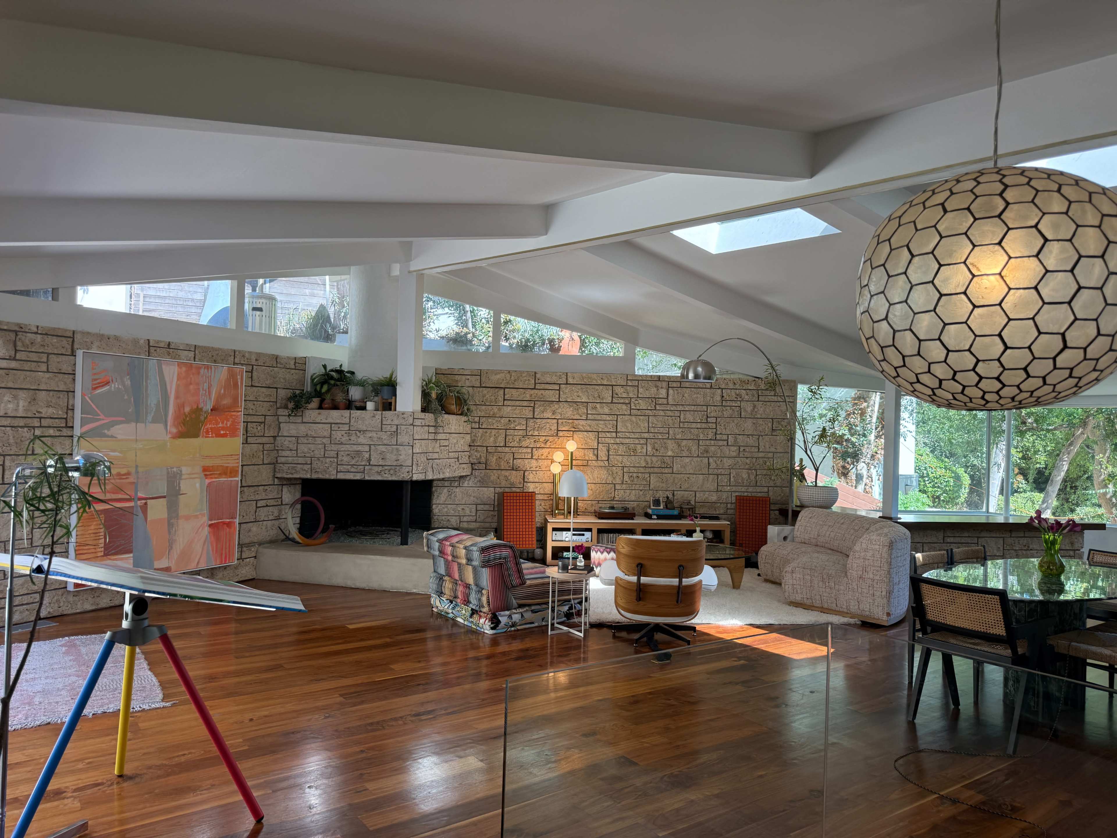 The image shows a spacious, modern living room with a stone fireplace, large windows, and a mix of furniture including a patterned couch, an armchair, and an easel displaying colorful artwork.