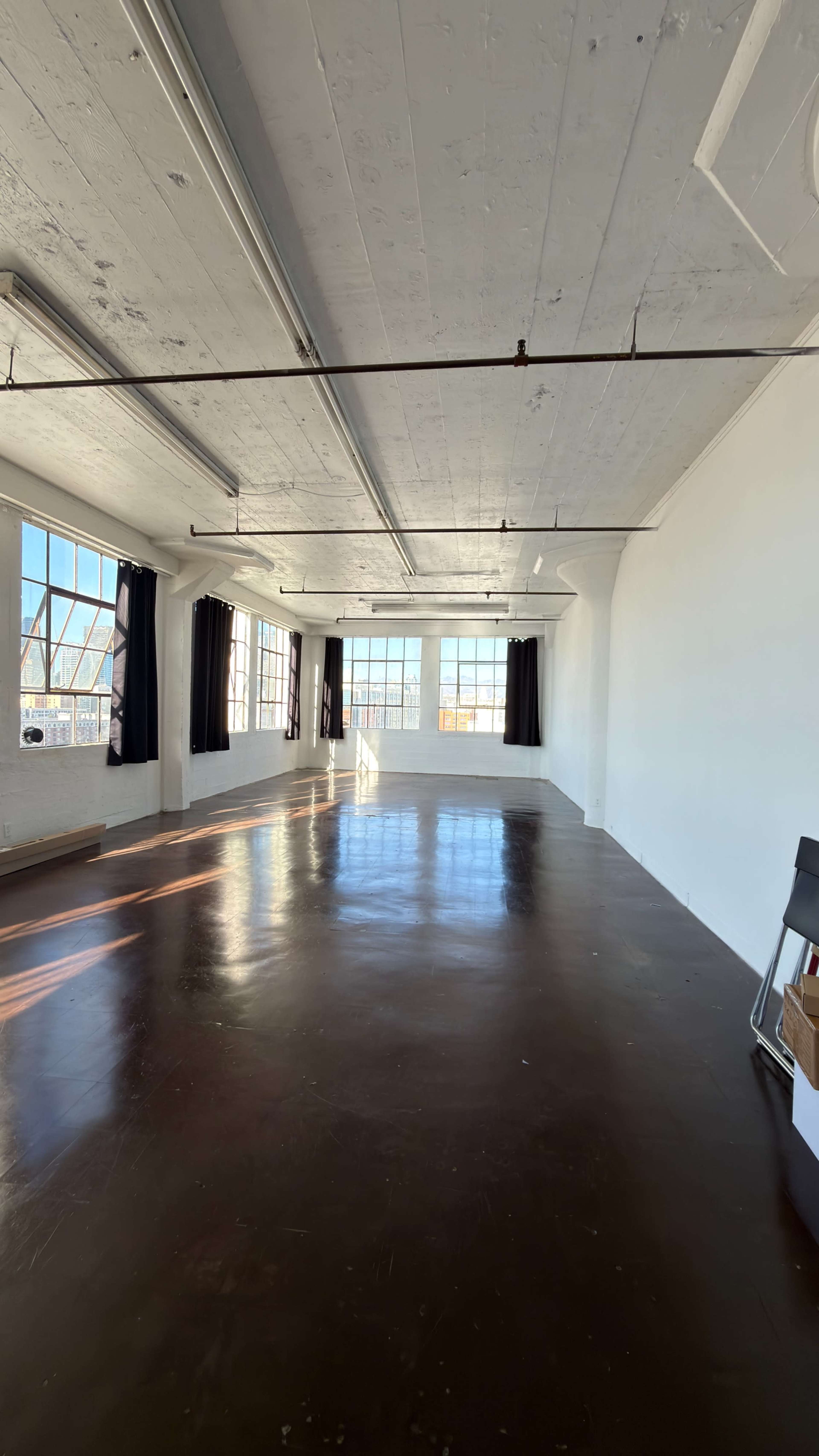 The image shows a spacious, well-lit room with large windows, a polished floor, and minimal furniture along the walls.