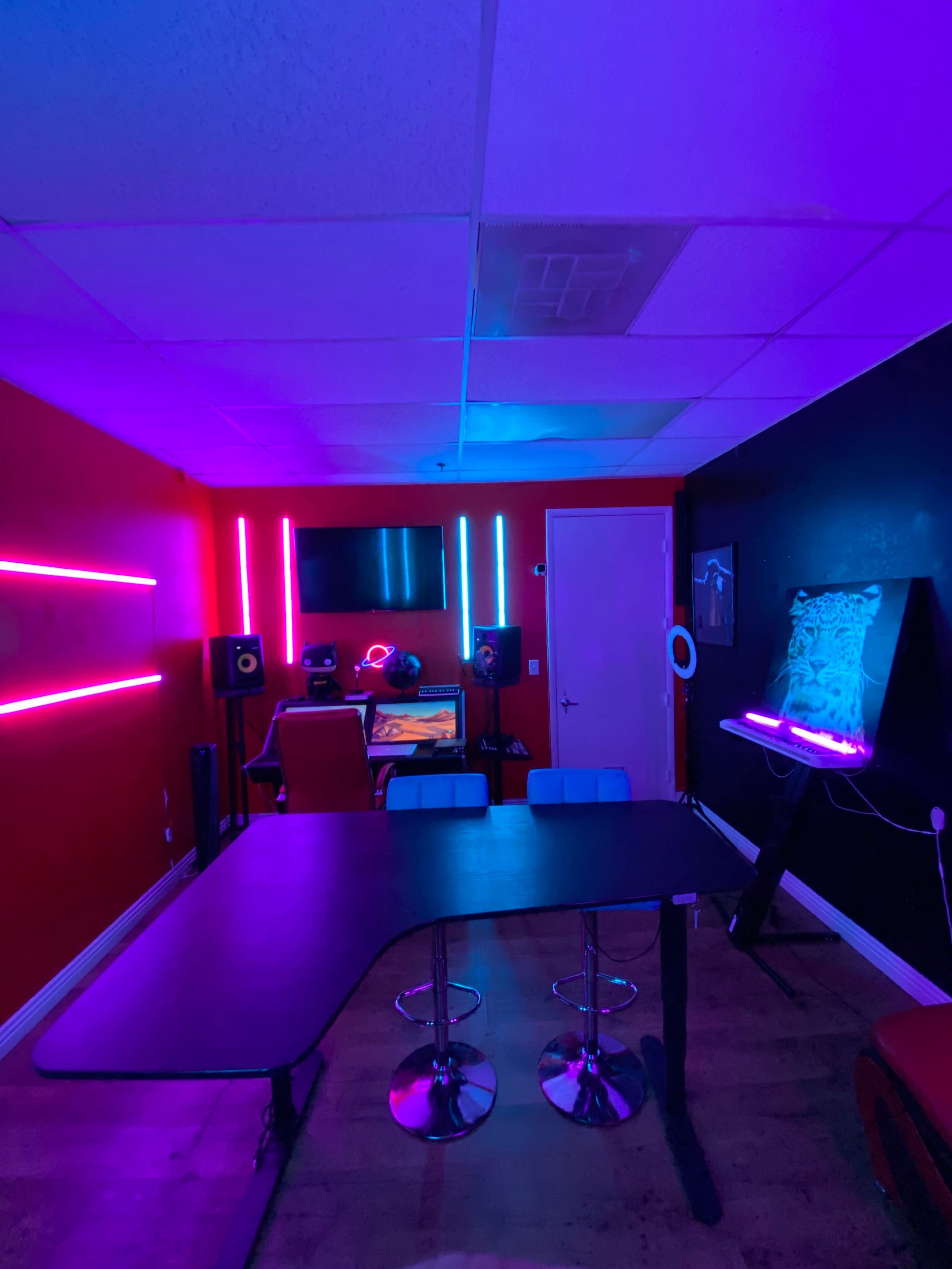A room with colorful LED lights features a desk, computer setup, and bar stools against a backdrop of red and black walls.