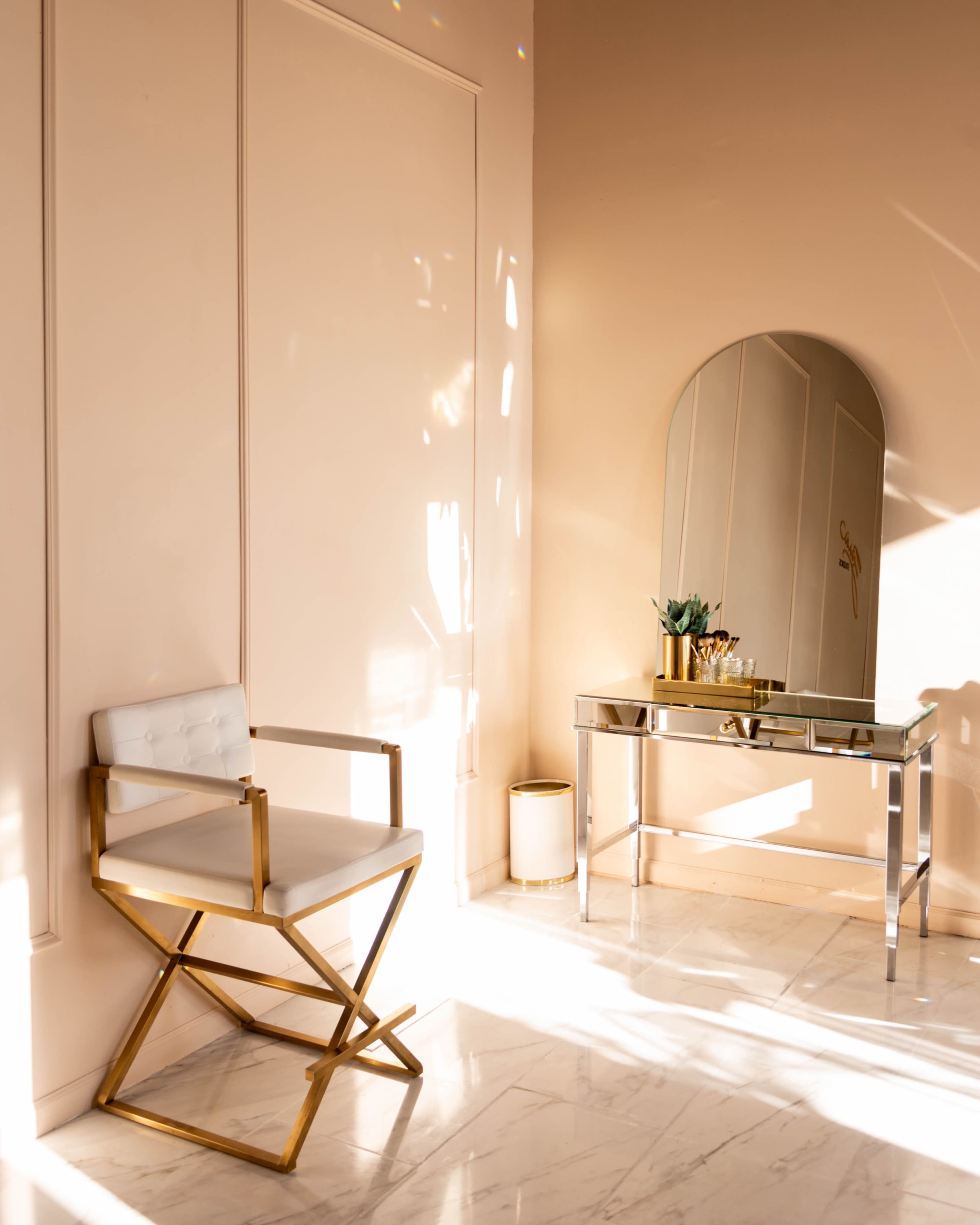 A stylish corner of a room featuring a white and gold chair beside a reflective console table with decorative items, all set against a light pink wall.