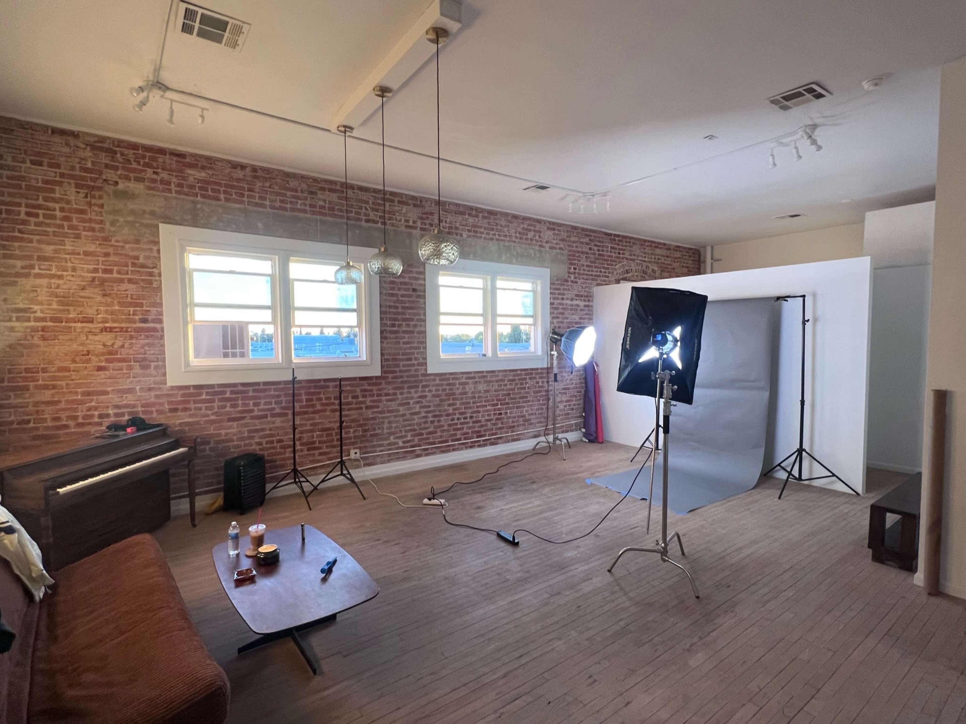 The image shows a spacious room with exposed brick walls, a piano, a coffee table, and a photography setup including a backdrop and lighting equipment.