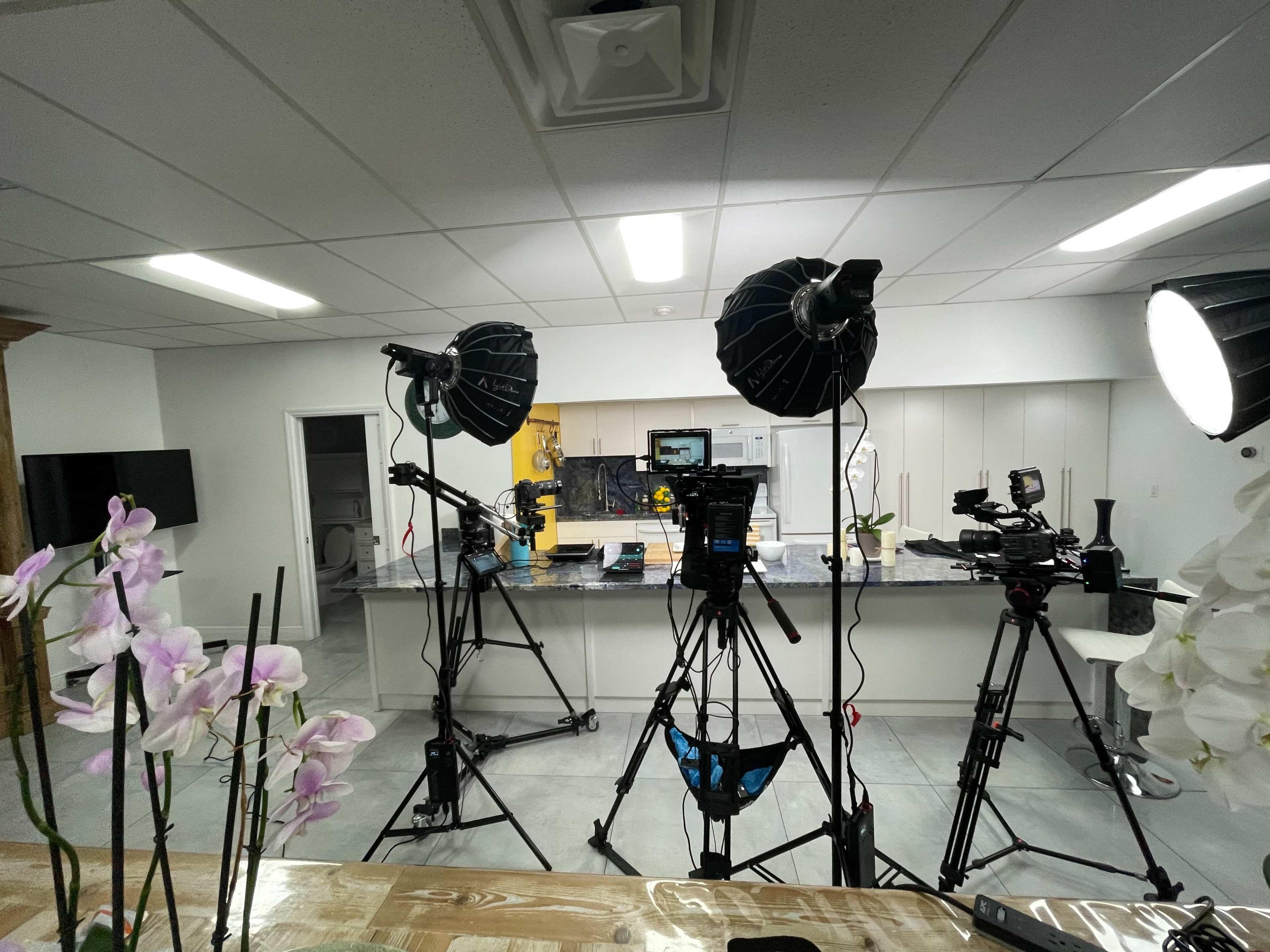 The image shows a well-lit kitchen set up for filming, featuring multiple cameras and lighting equipment in front of a counter.