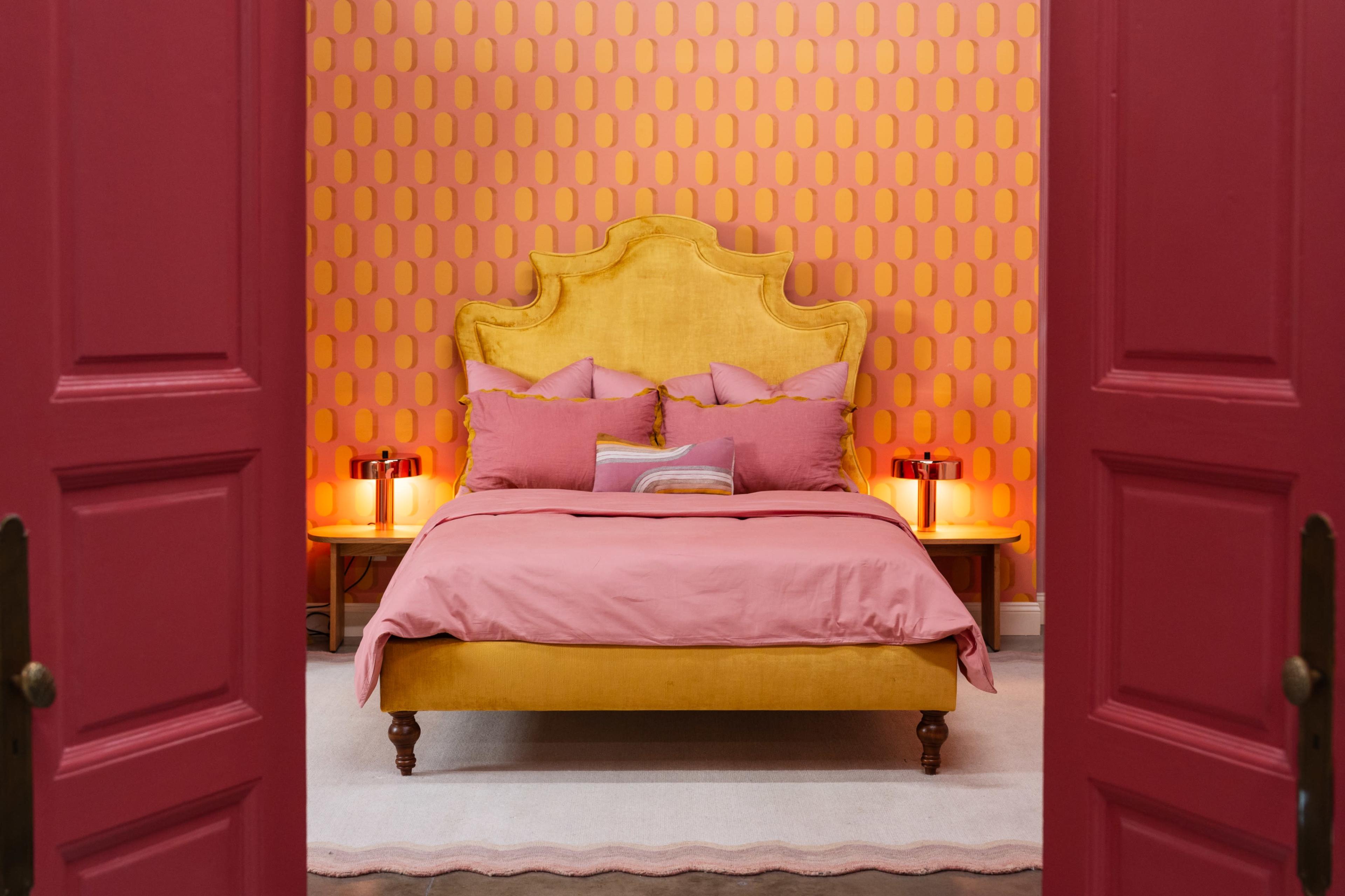 The image shows a brightly colored bedroom featuring a yellow bed with pink bedding, surrounded by pink patterned walls and illuminated by glowing lamps on either side of the bed.
