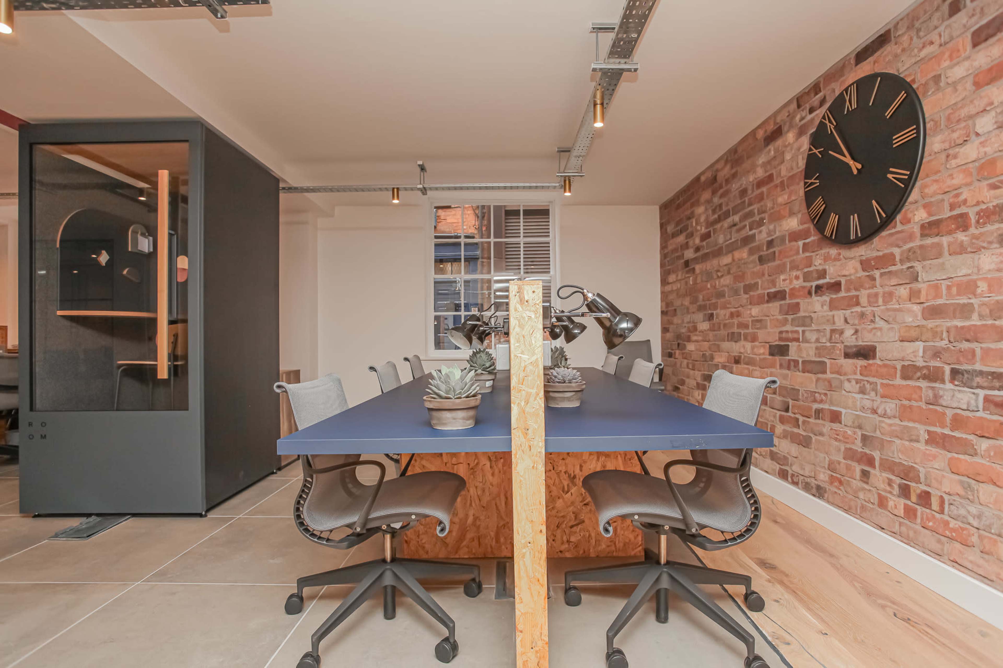 The image shows a modern office meeting room featuring a long blue table with rolling chairs, a brick wall, and a clock, along with a glass-enclosed workspace in the background.