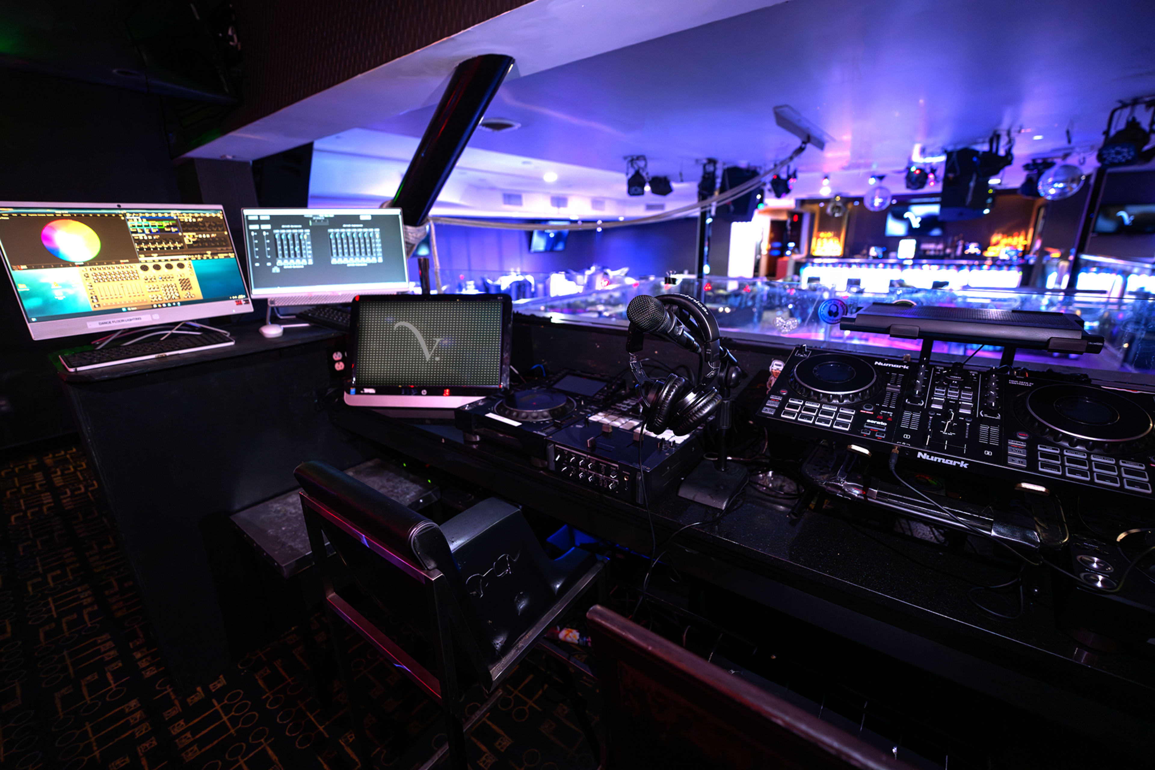 The image shows a DJ booth in a club, featuring multiple screens and equipment, including mixers and turntables.