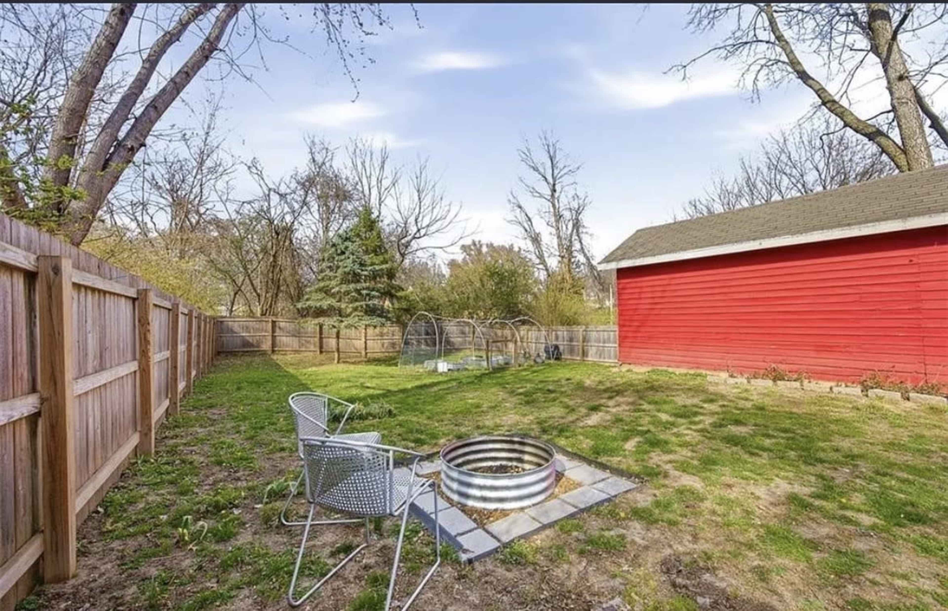 The image shows a backyard with a red shed, a fire pit on stone tiles, two white chairs, and a garden area surrounded by a wooden fence.