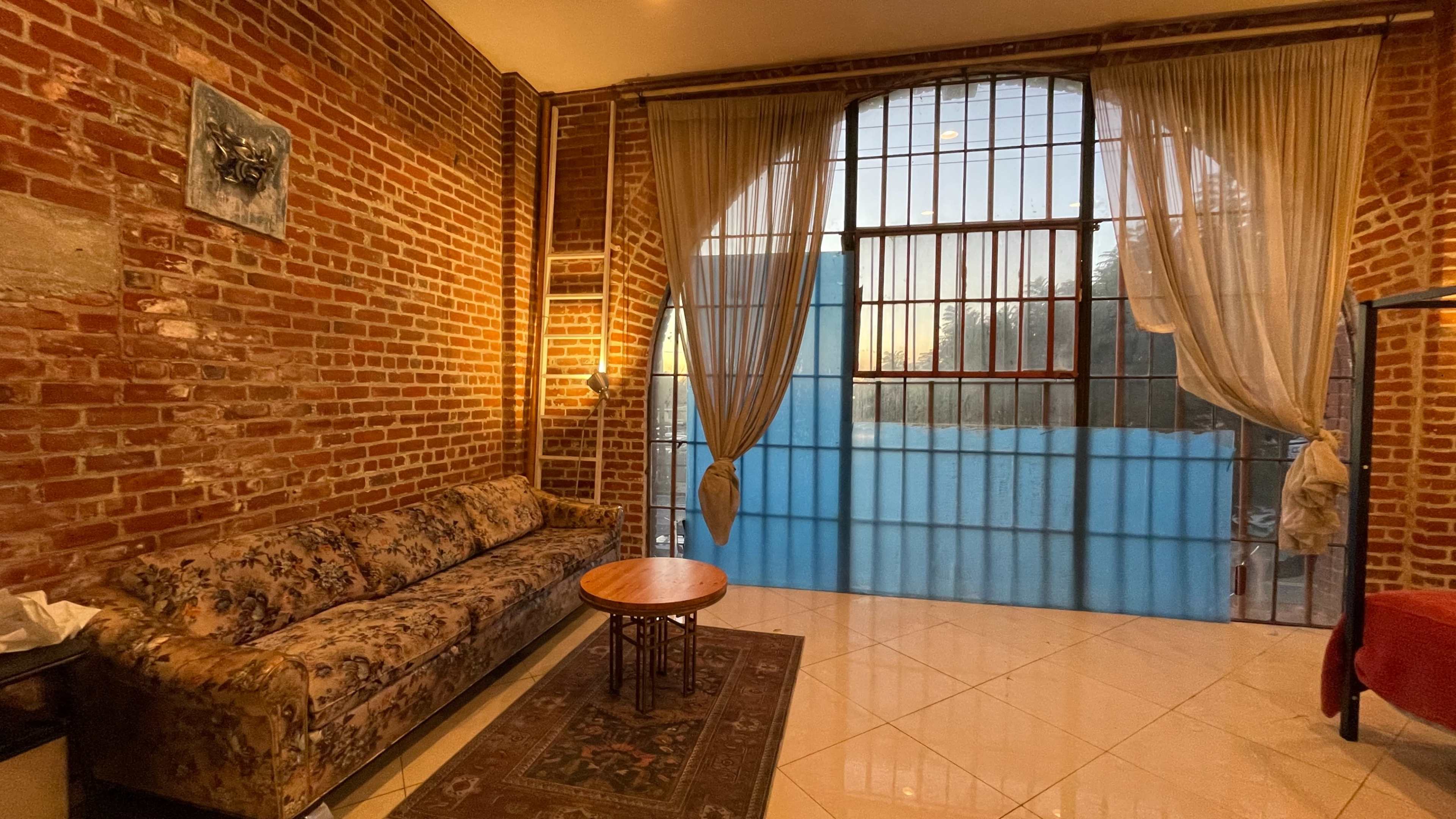 The image shows a cozy interior featuring a floral-patterned sofa, a small round table, and large windows covered by sheer curtains, with exposed brick walls creating a warm atmosphere.