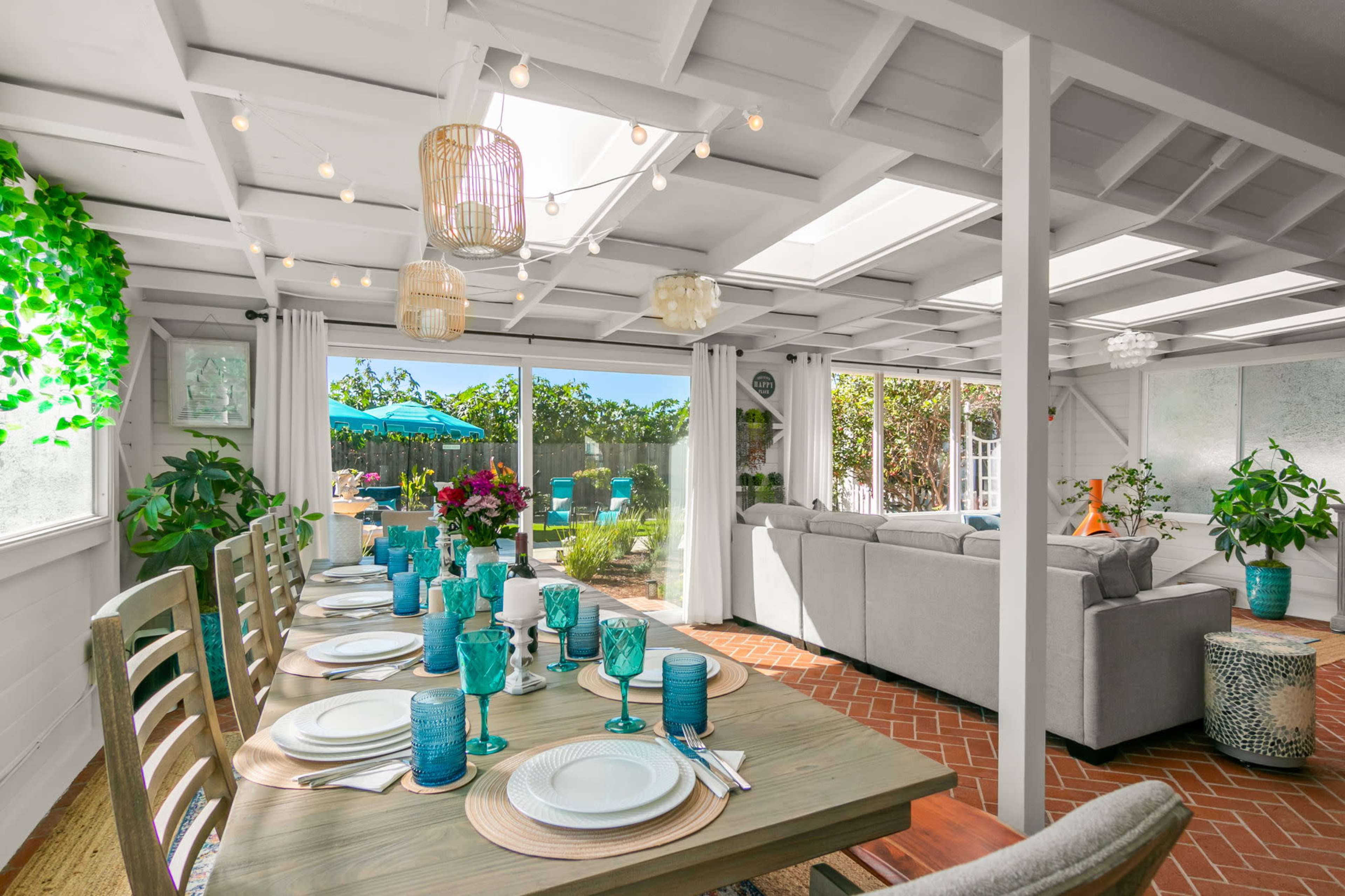 A dining area features a long wooden table set with turquoise glasses and white plates, surrounded by plants, and opens up to a bright outdoor space.