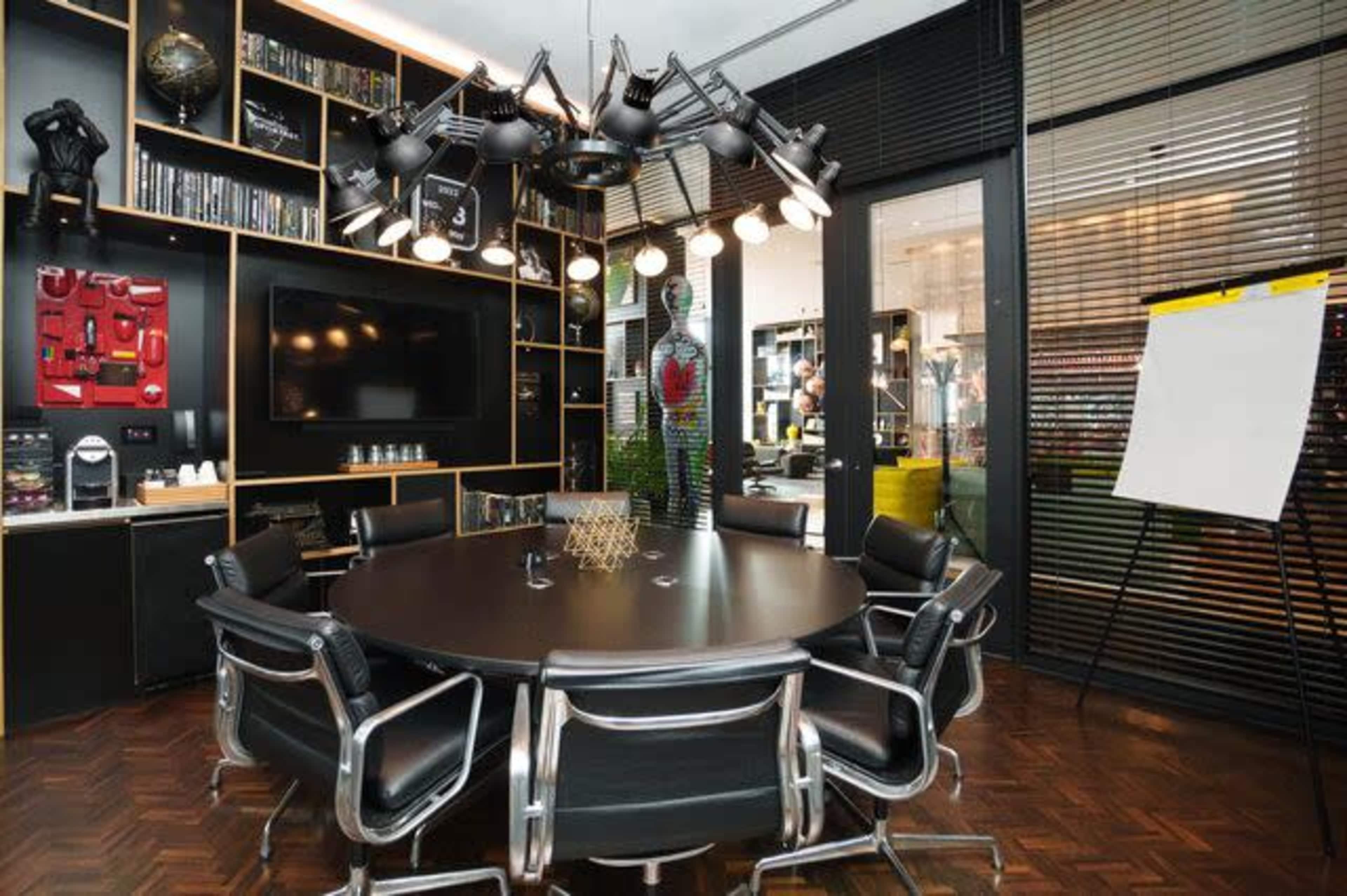 A modern conference room features a round table surrounded by sleek black chairs, a large TV, and wall-mounted bookshelves filled with various items.