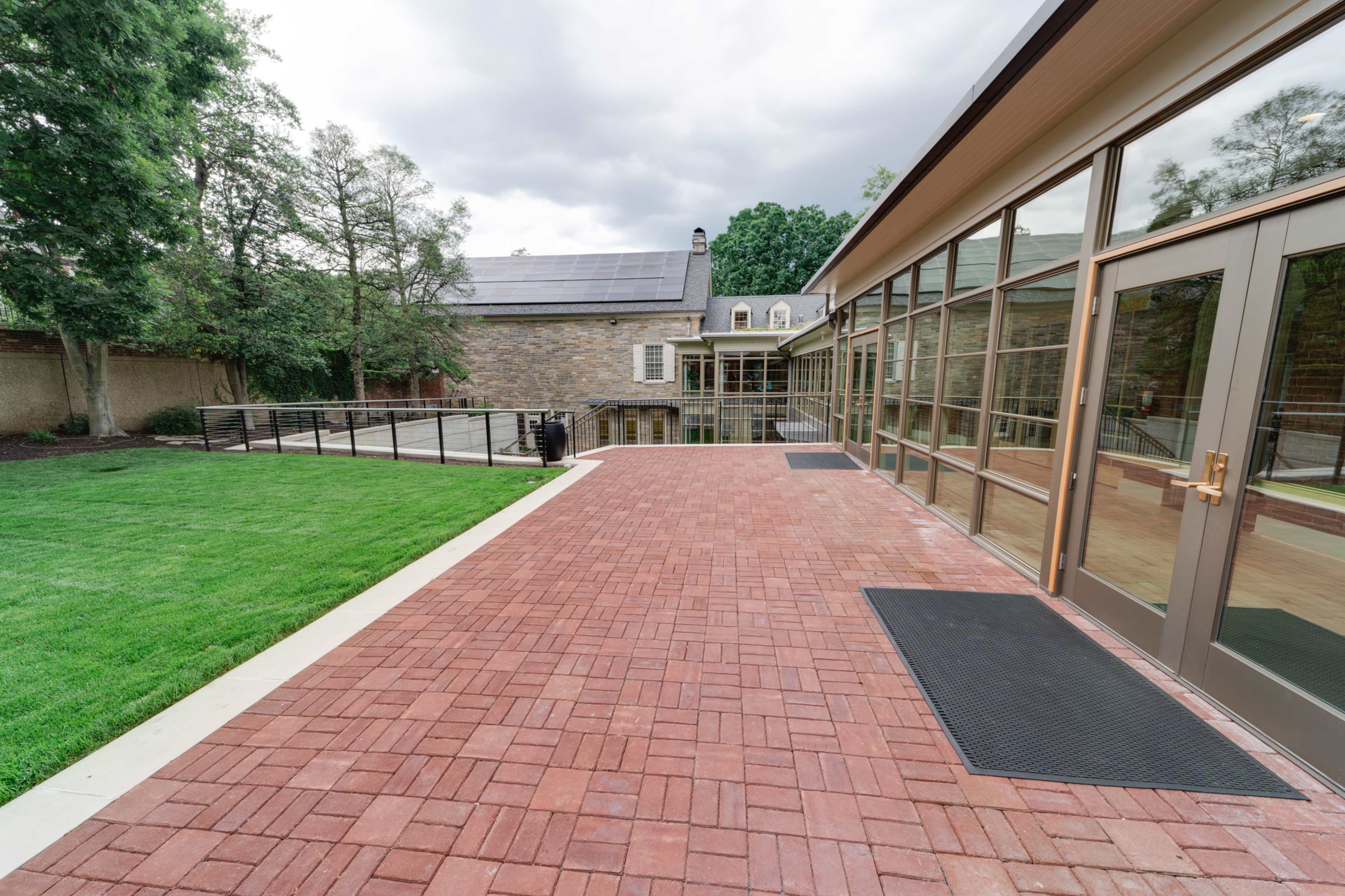 A brick patio area adjacent to a modern glass building and a stone structure, with green grass and trees in the background under a cloudy sky.