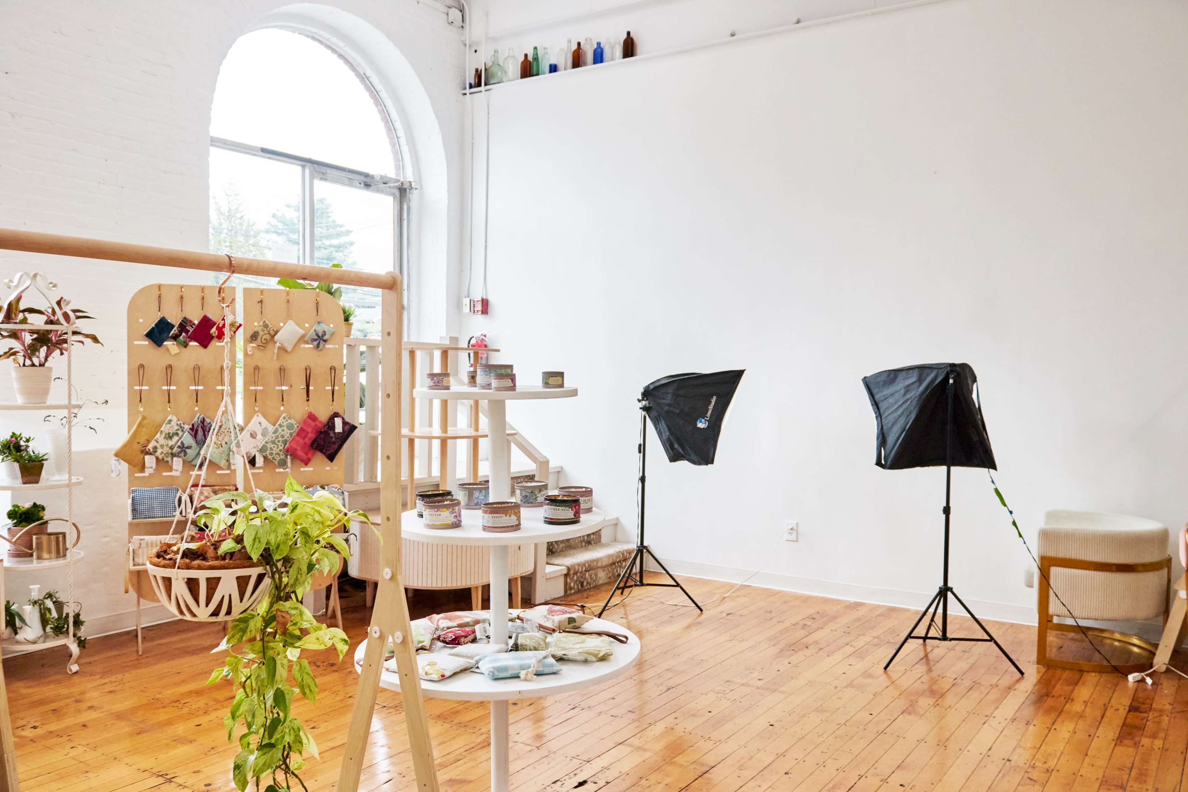 A bright, airy room with wooden floors, featuring a display of plants, handmade items on tables, and photography lights set up in the corners.