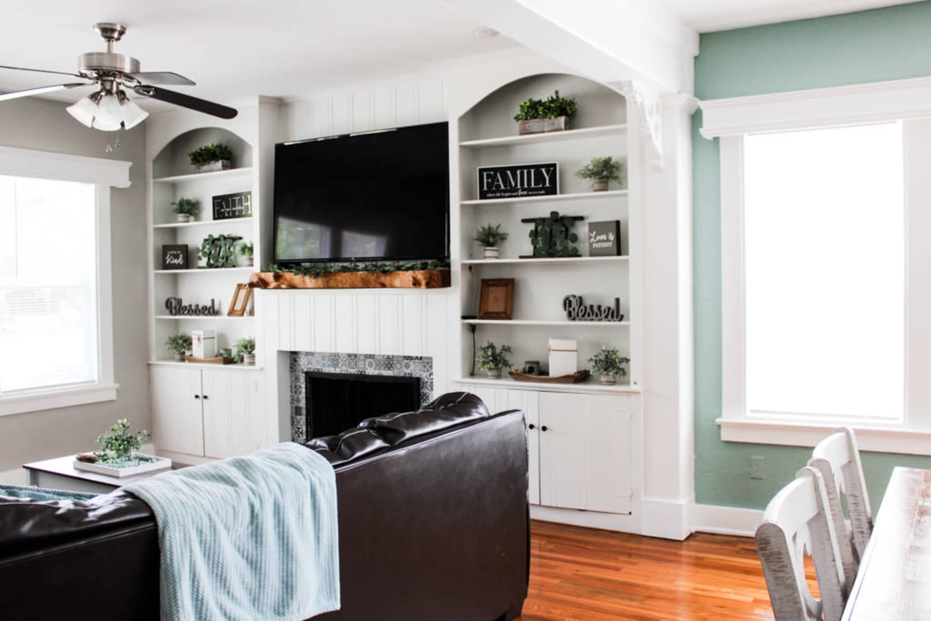 A living room with a sectional sofa, a television mounted on built-in shelves, and a light-colored dining table nearby.