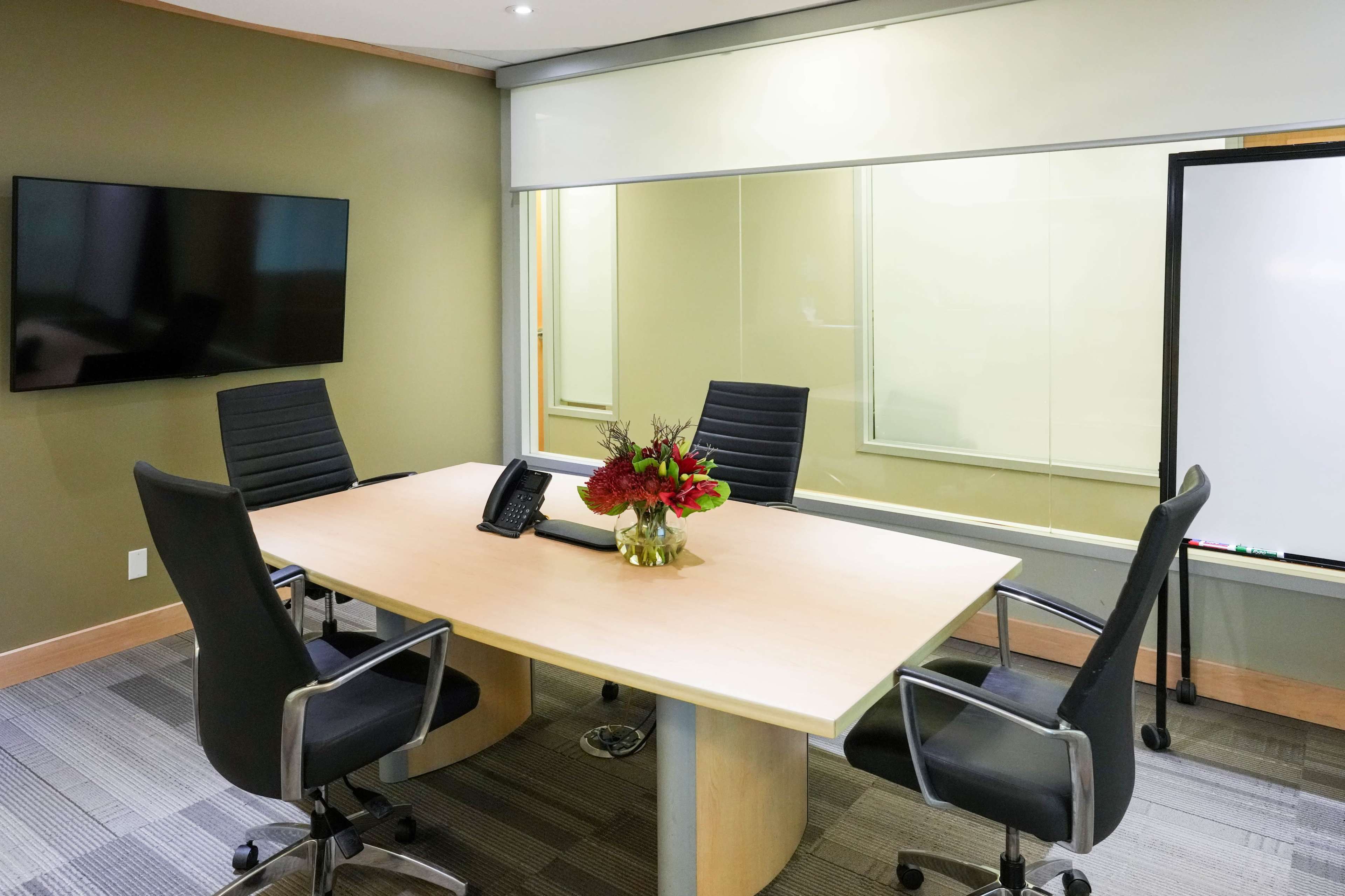A meeting room features a round wooden table surrounded by five black chairs, a television mounted on the wall, and a bouquet of flowers in the center.