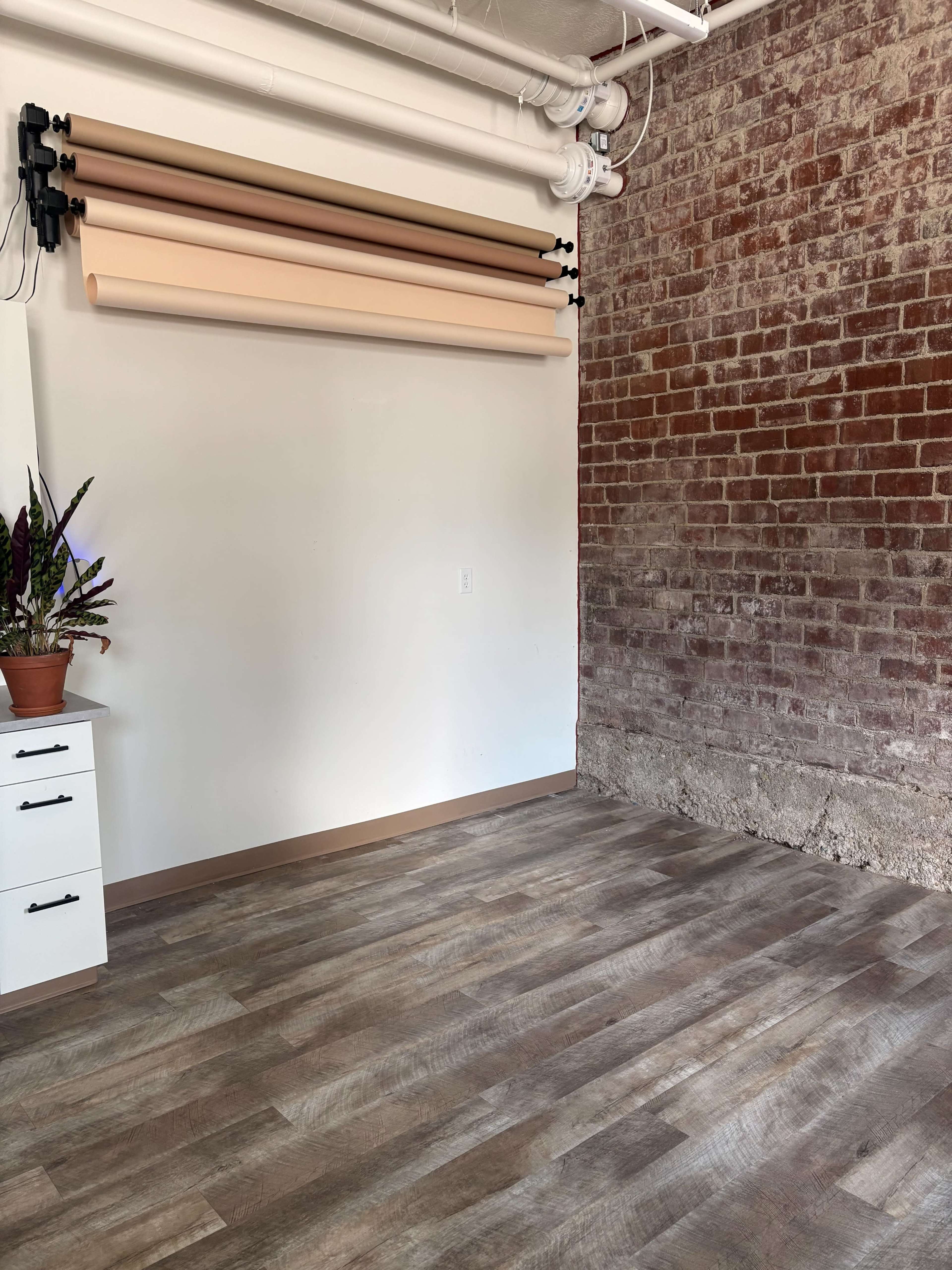 The image shows a spacious room with a section of exposed brick wall, hardwood flooring, and a wall-mounted backdrop holder with various colored paper rolls.
