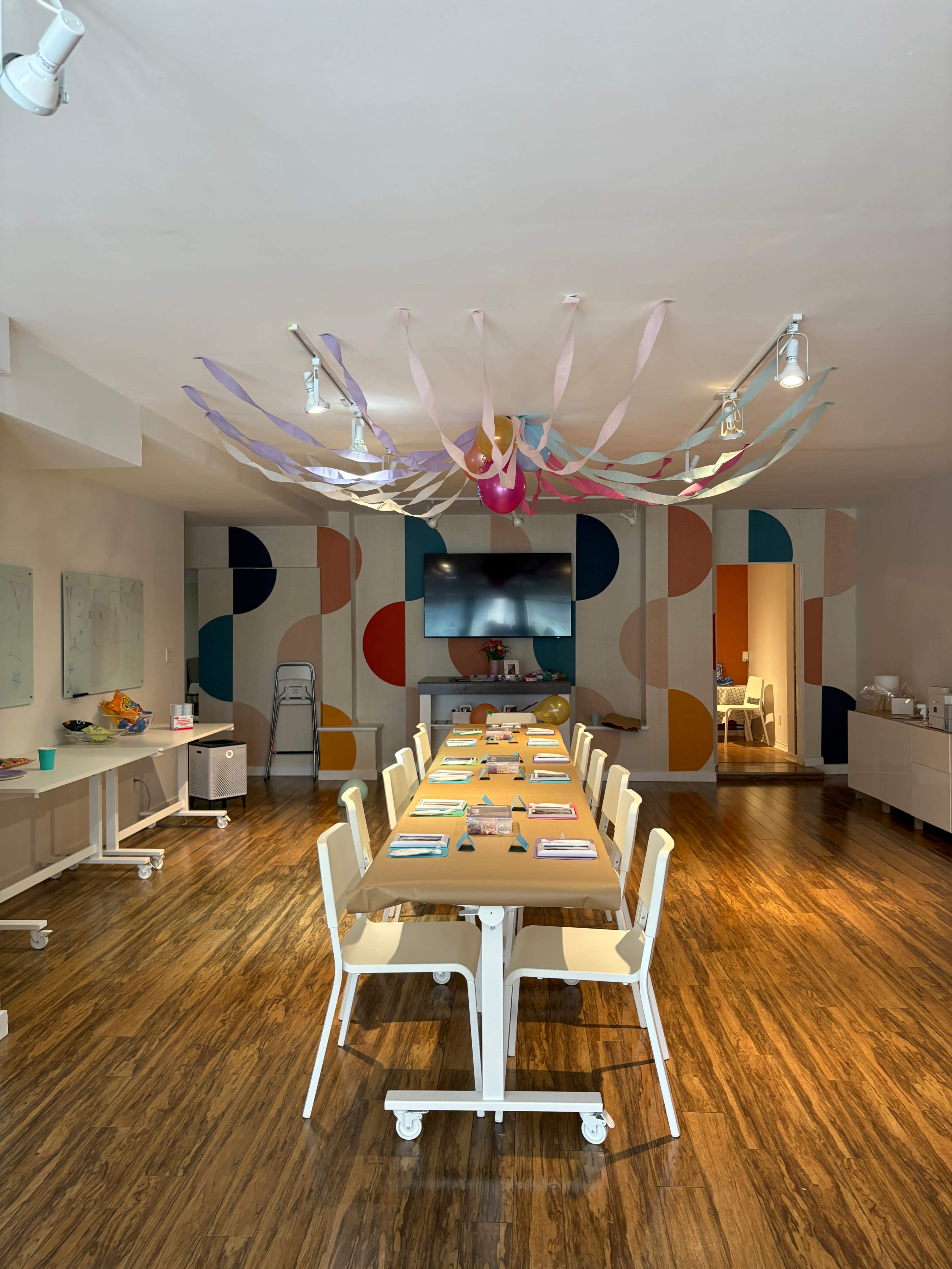 A long table is set with colorful materials in a modern meeting room featuring abstract wall decorations and streaming lights overhead.
