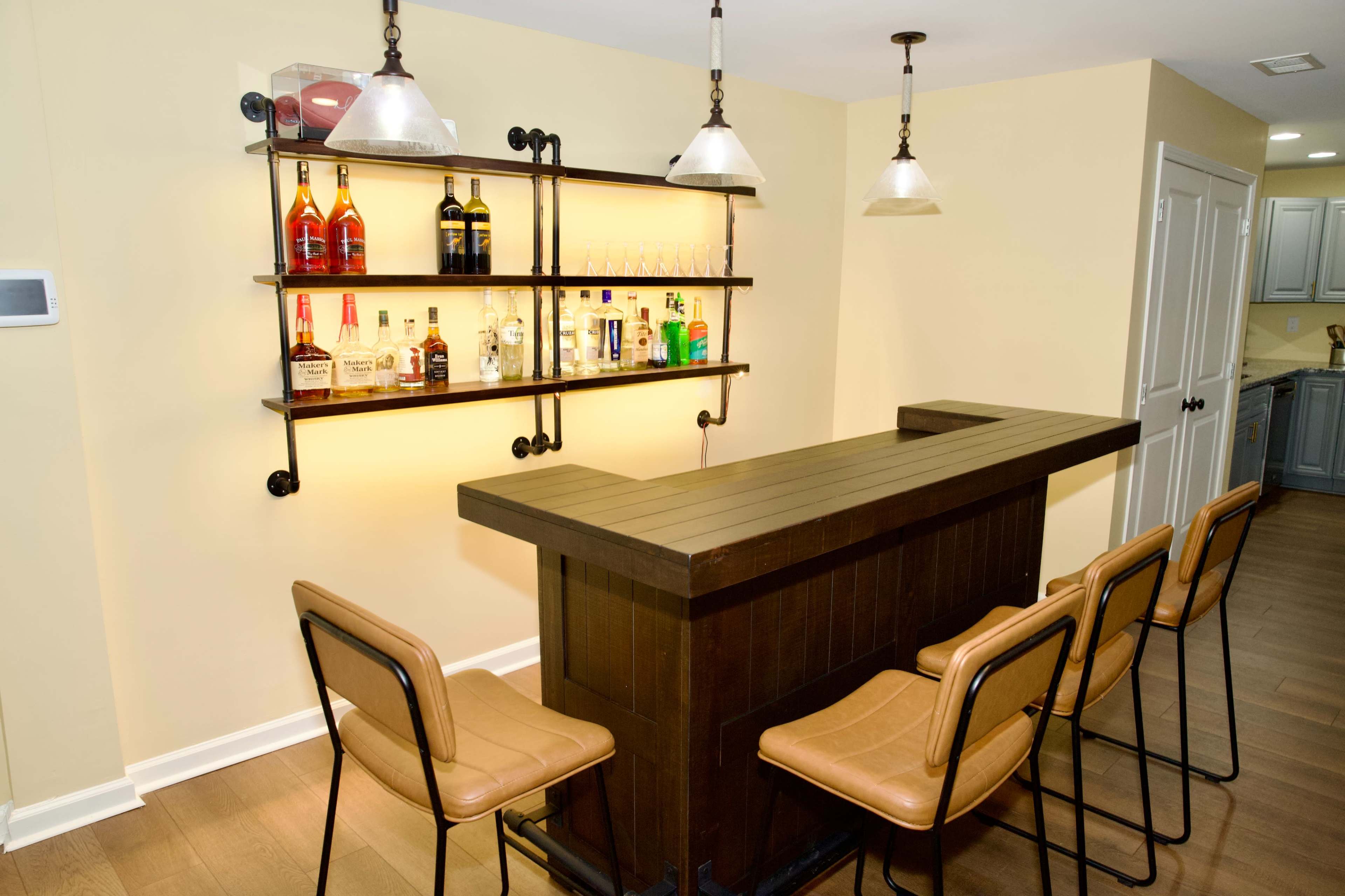 The image shows a modern home bar with a wooden counter, six metal-framed stools, and shelves displaying various liquor bottles.
