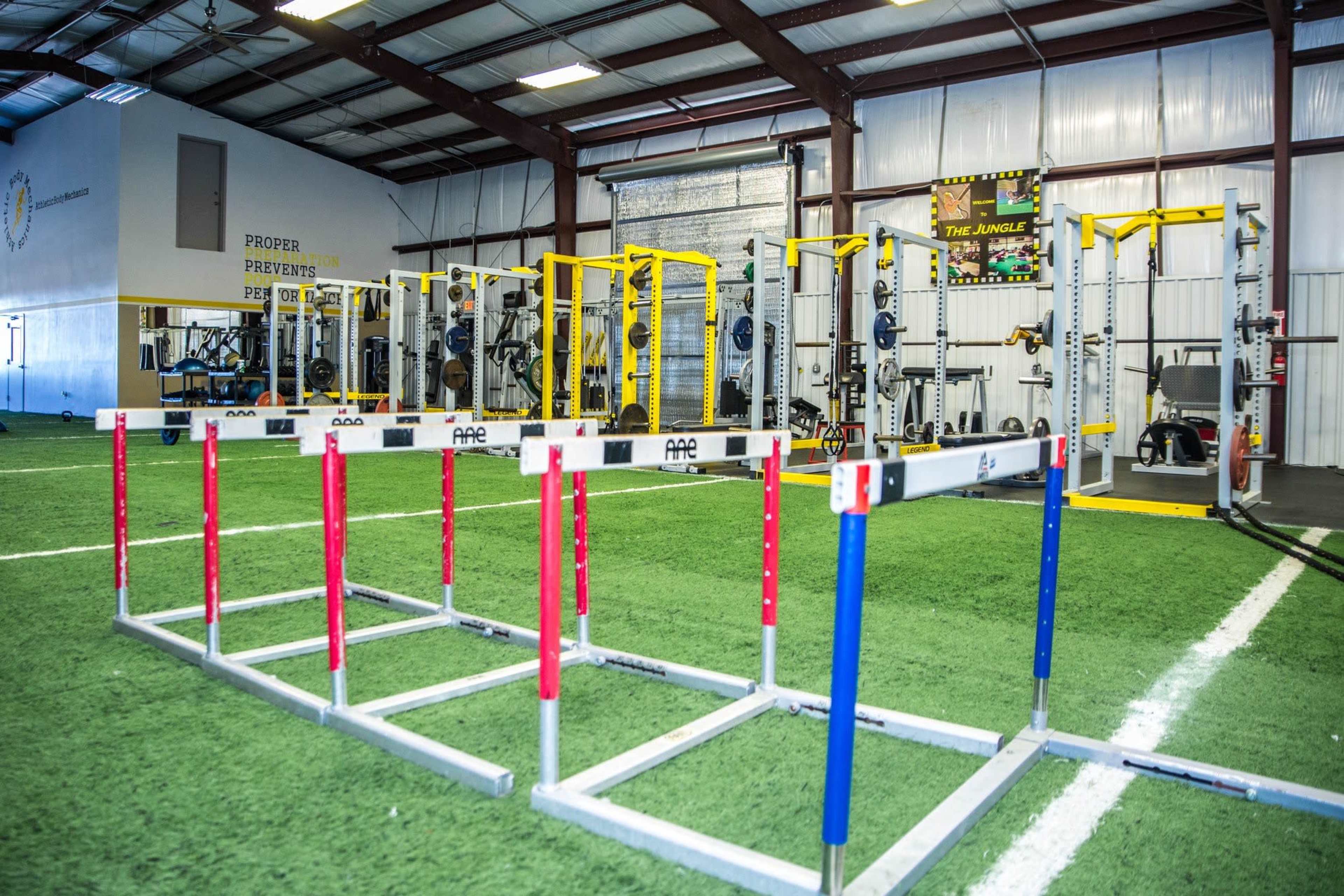 A training facility with agility hurdles positioned in front of a gym area equipped with weightlifting racks and exercise machines.