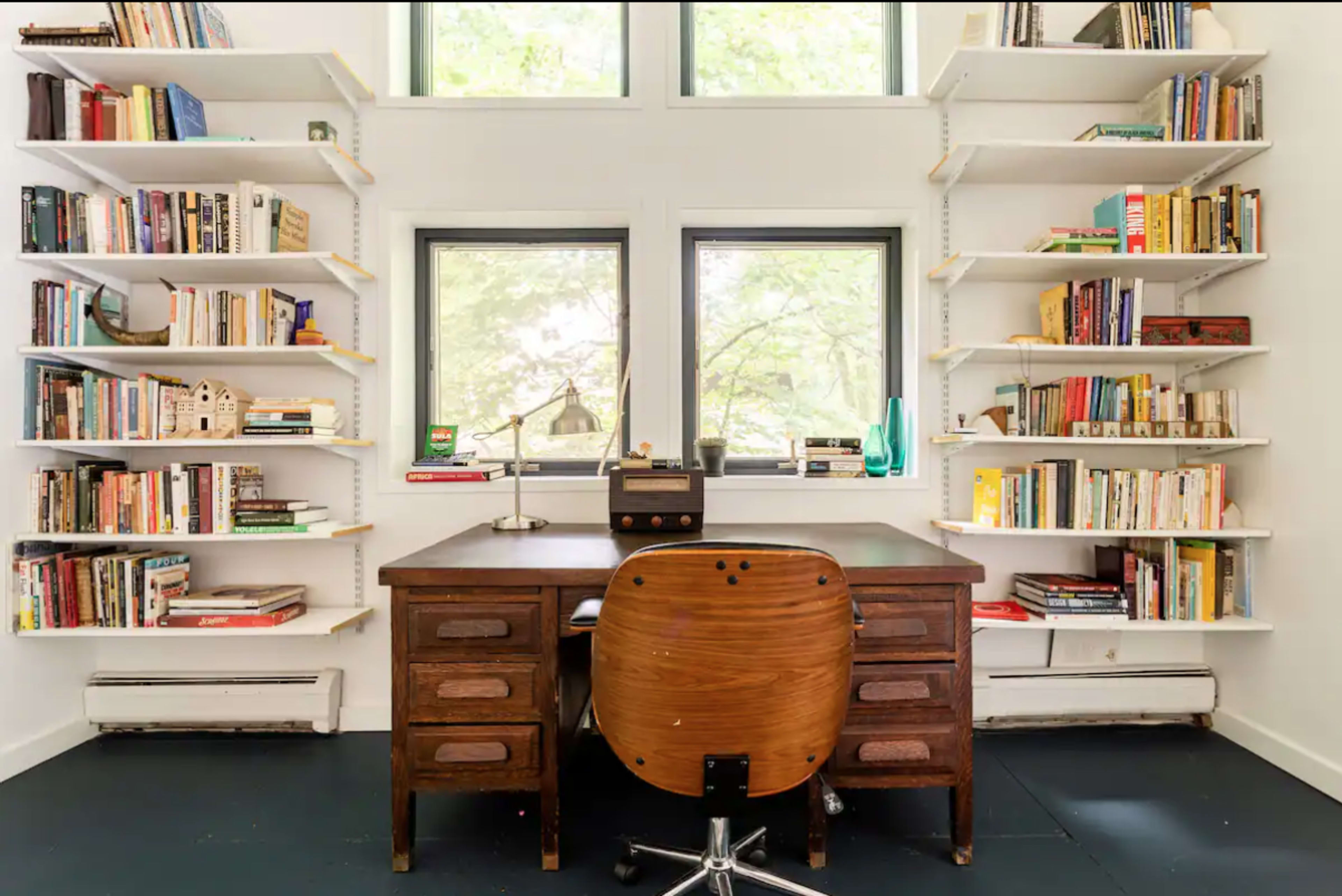 A wooden desk with a chair sits in the center of a room surrounded by bookshelves filled with various books.