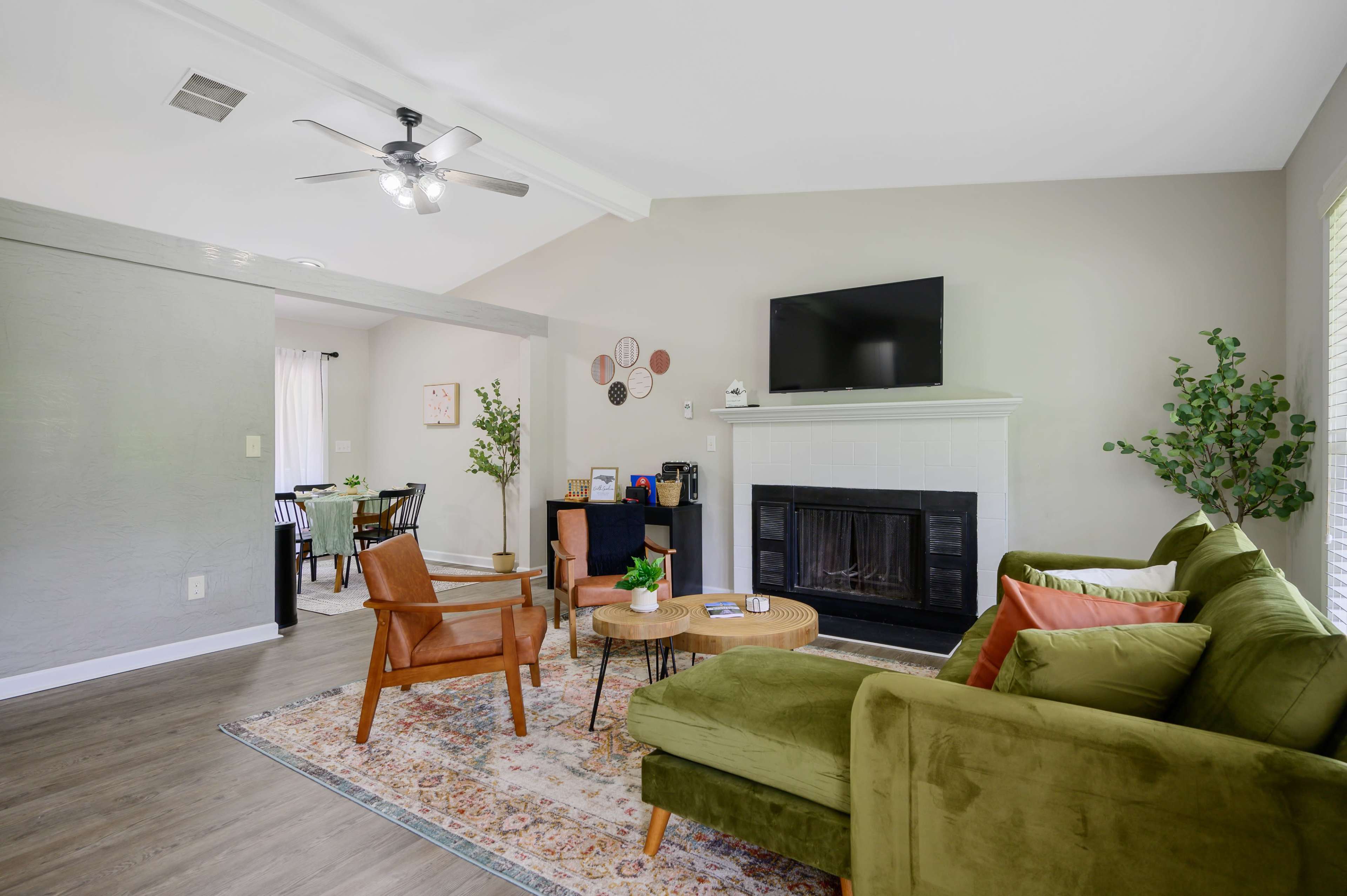 A living room features a green sofa, a wooden coffee table, and a flat-screen TV mounted above a white fireplace, with plants and decorative items visible.