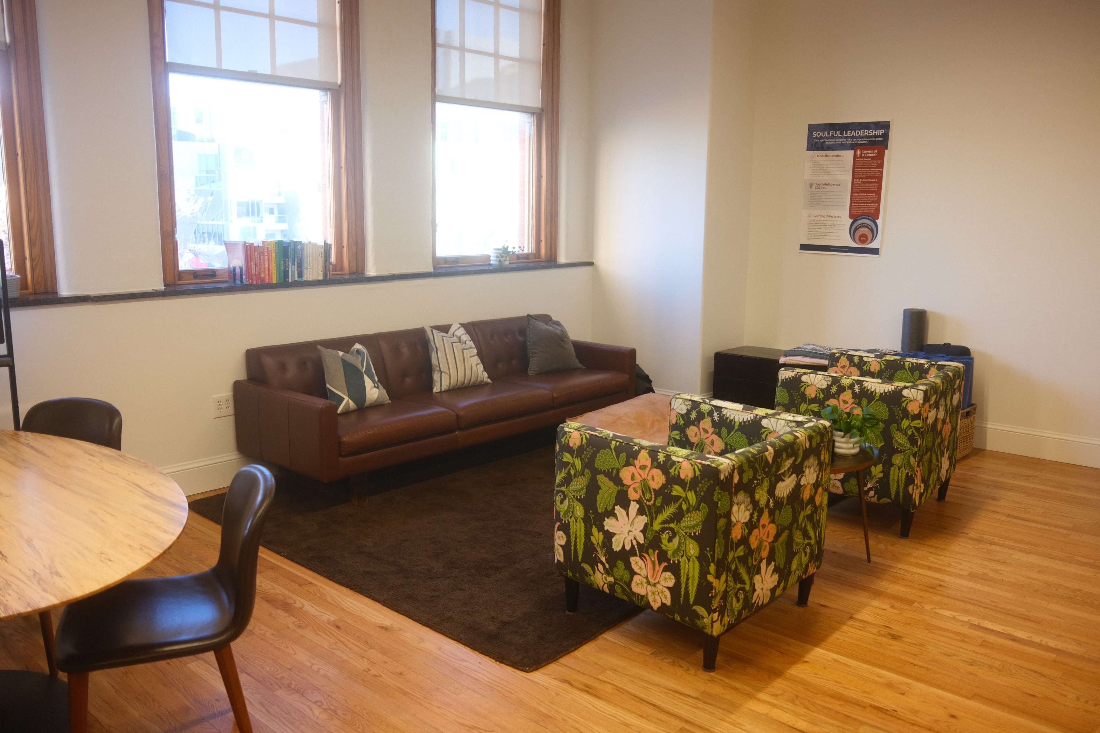 The image shows a bright room featuring a brown leather sofa, two floral-patterned chairs, a round wooden table, and large windows letting in natural light.