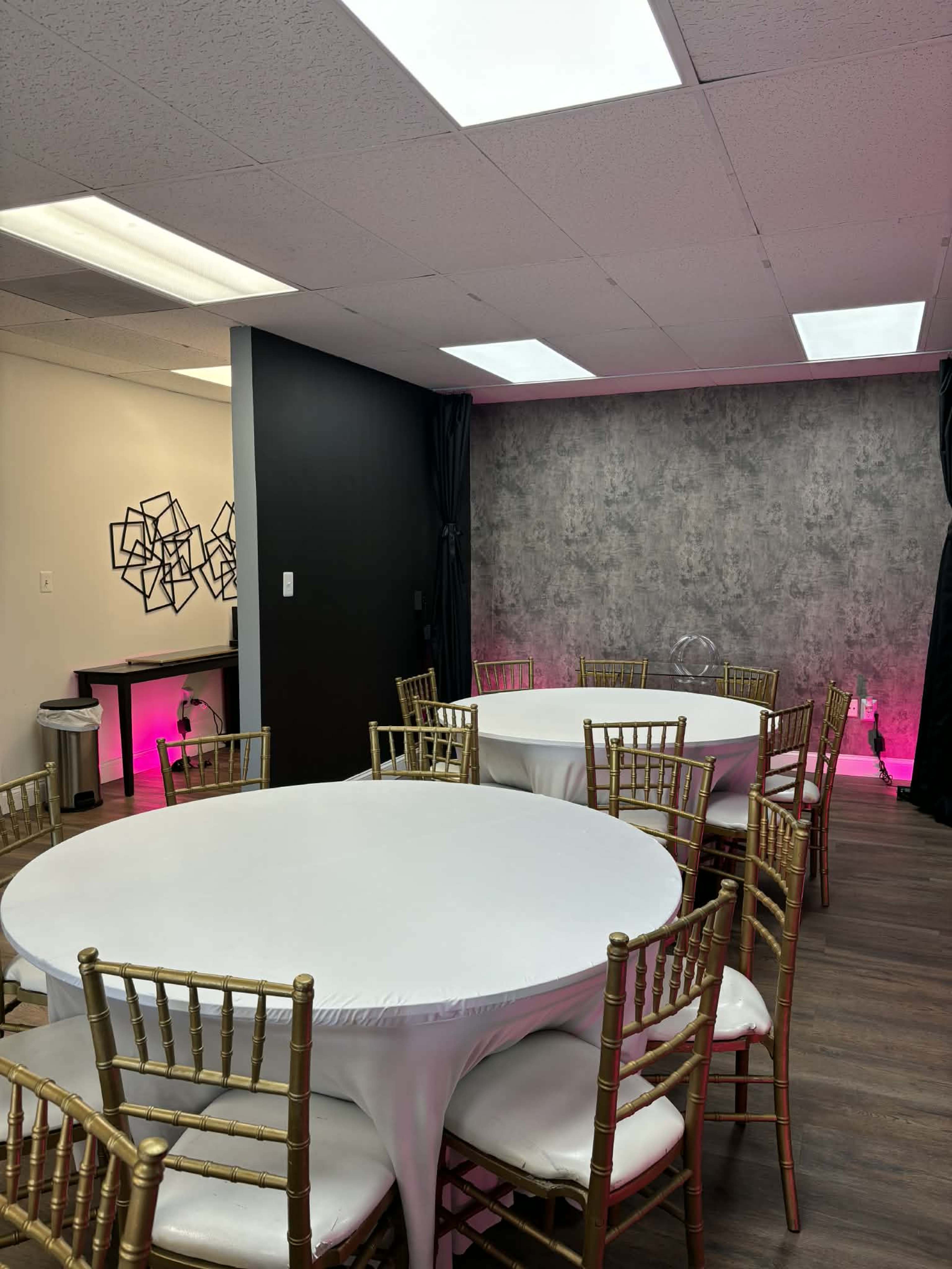 The image shows a room with several round tables surrounded by gold chairs, a gray wall, and pink lighting in the background.