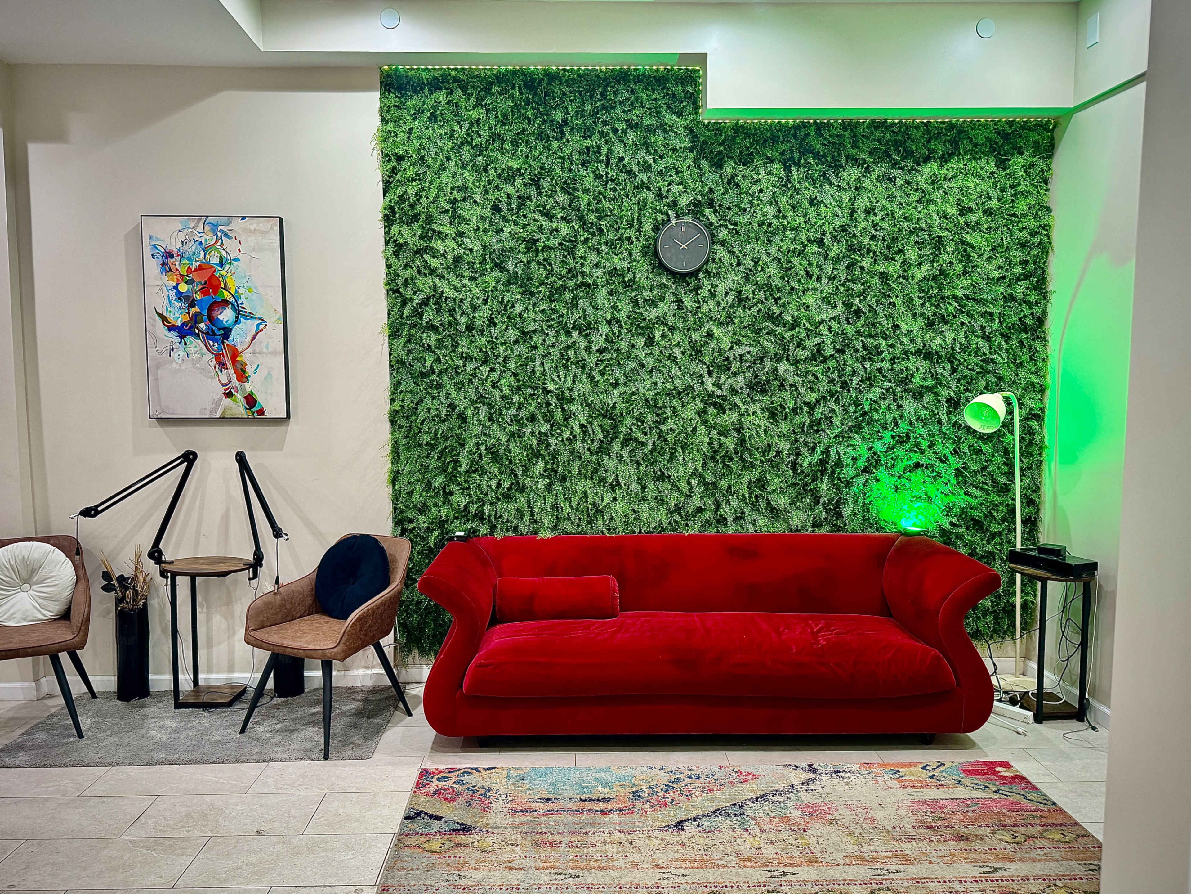 A red couch is placed in front of a vibrant green wall adorned with a clock, accompanied by two chairs and a lamp in a well-lit room.