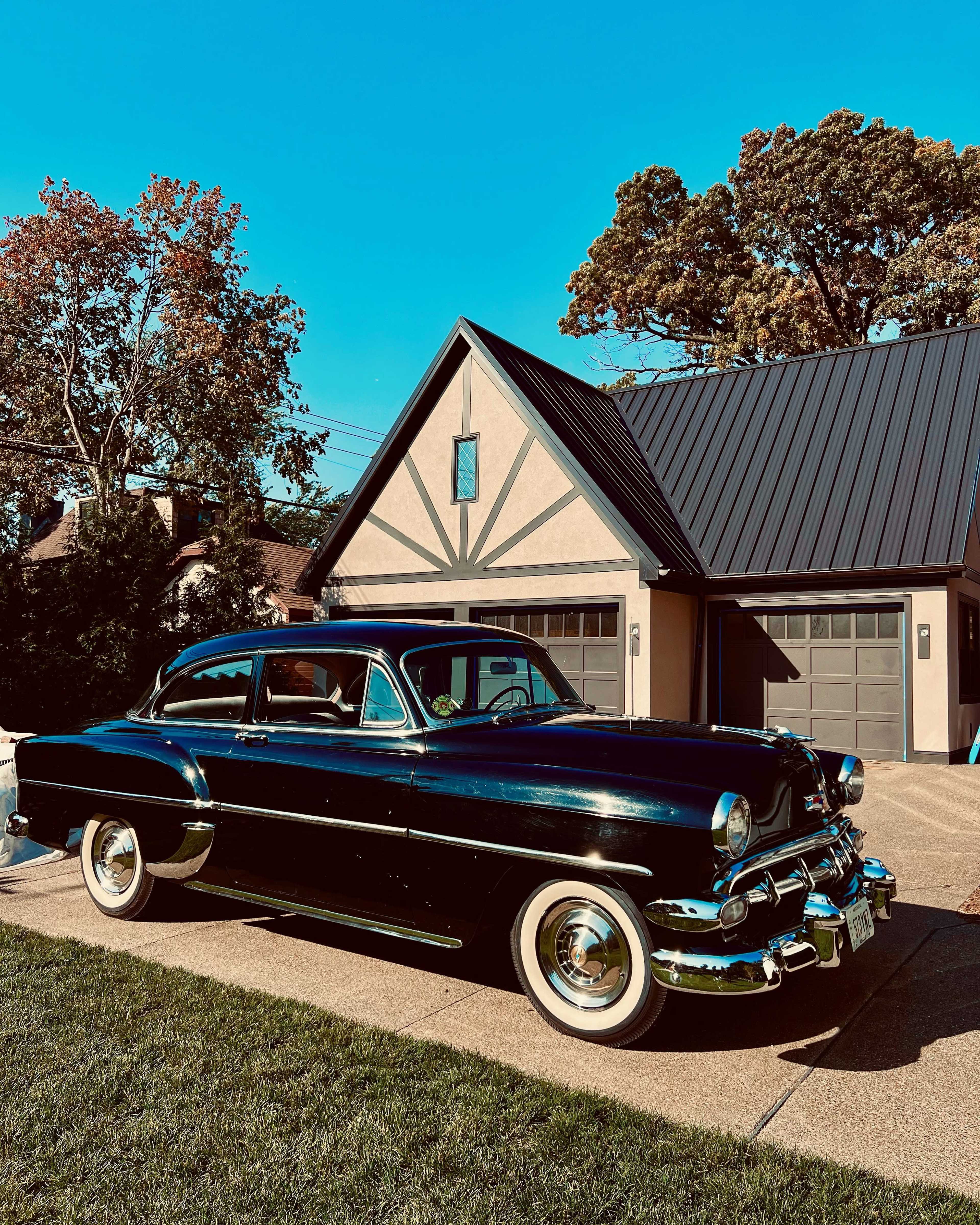 A classic black car is parked in front of a modern house with a steeply pitched roof and twin garage doors.